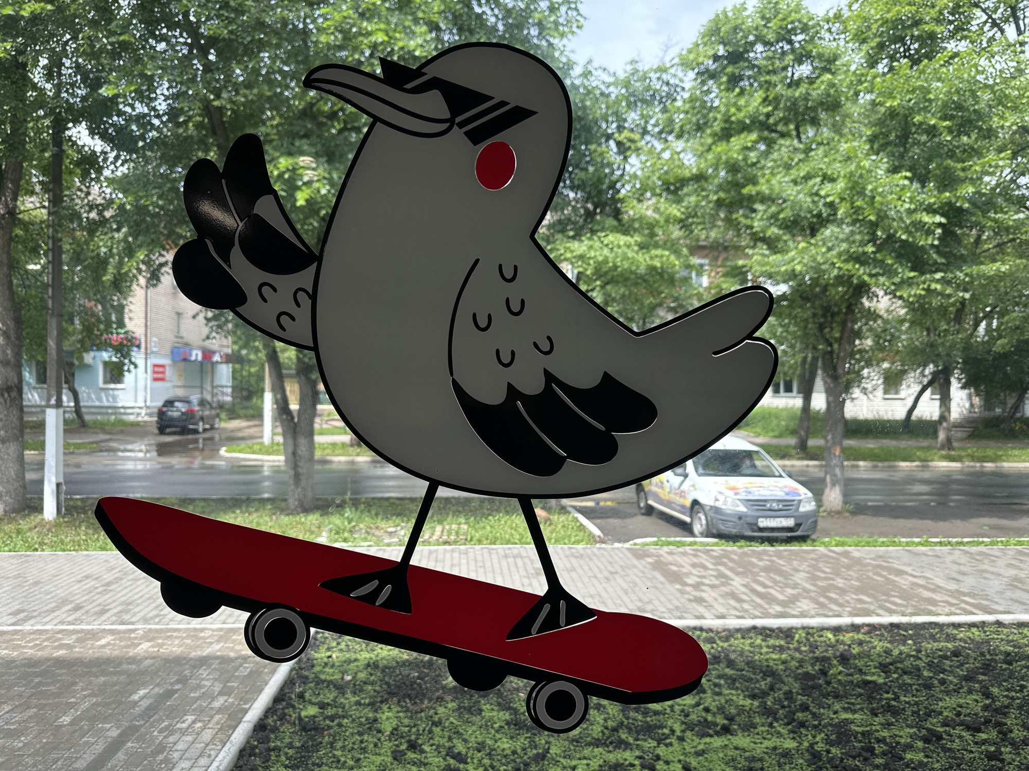 A sticker of a cartoon bird with sunglasses and a red eye is stuck to a window overlooking a street in Chaikovsky, Russia. The bird is gray with black accents and is depicted riding a red skateboard. In the background, trees line a wet sidewalk and a street with parked cars. The overall scene suggests an urban, possibly rainy day.
