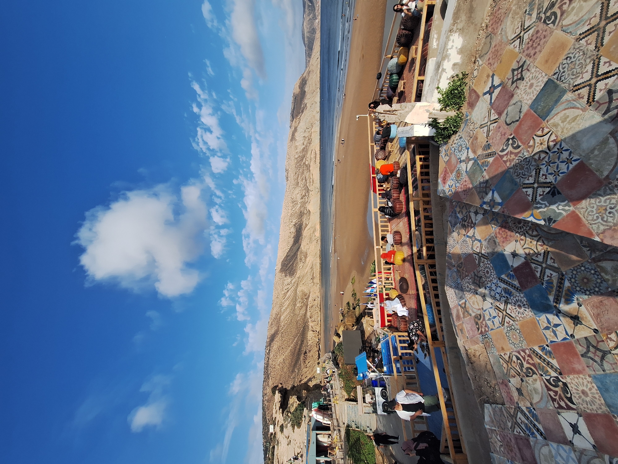 This is a panoramic, high-angle shot of a seaside cafe in Imsouane, Morocco, on a clear day with a bright blue sky and scattered white clouds. In the foreground, a tiled patio with a colorful, geometric mosaic pattern leads down towards a wooden deck. The deck is furnished with low seating areas, including cushioned poufs and floor mats, occupied by several individuals. Some people are interacting, while others are seated individually, observing the surroundings. Beyond the deck, a wide, sandy beach stretches to meet the calm, blue ocean. In the distance, a rugged, arid mountain range forms the backdrop. Along the edge of the beach and the foothills, several small buildings and structures are visible, some appearing to be part of the cafe's infrastructure, possibly housing surf shops or changing rooms. A few people can be seen walking on the beach and in the shallow water. The overall scene suggests a relaxed atmosphere at a popular coastal destination.