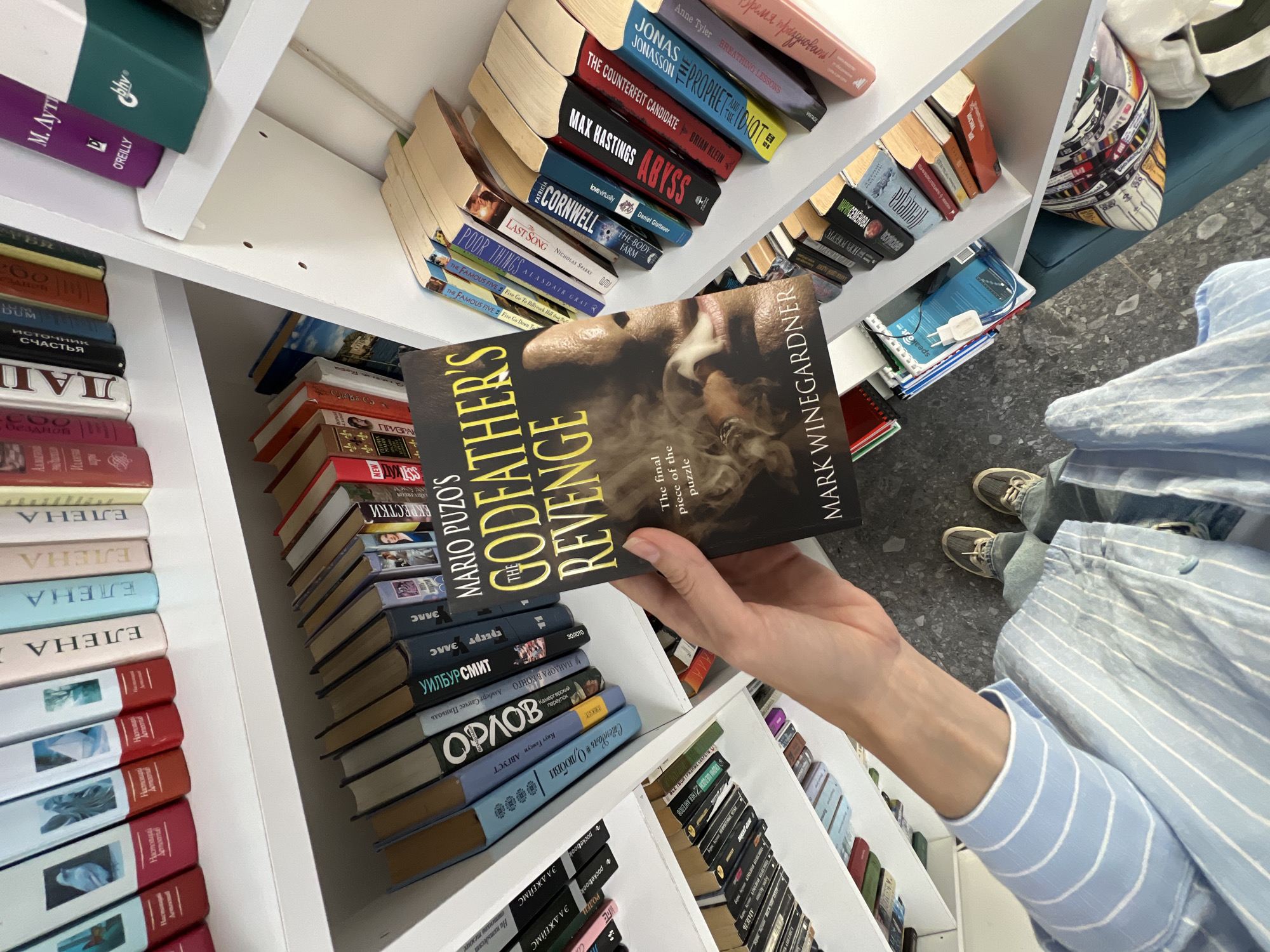 An individual holds the book "The Godfather's Revenge" by Mario Puzo and Mark Winegardner in front of a bookshelf. The shelf is filled with books arranged vertically, showing their spines. The titles and authors are visible, including books in Russian, English, and possibly other languages. The person is wearing a blue and white striped shirt and jeans, and their shoes are visible on the tiled floor. The photo is taken from above, looking down at the person and the bookshelf, with a pillow in the upper right corner. The location is within a bookstore or library. Bar, Montenegro is not visible in the image.
