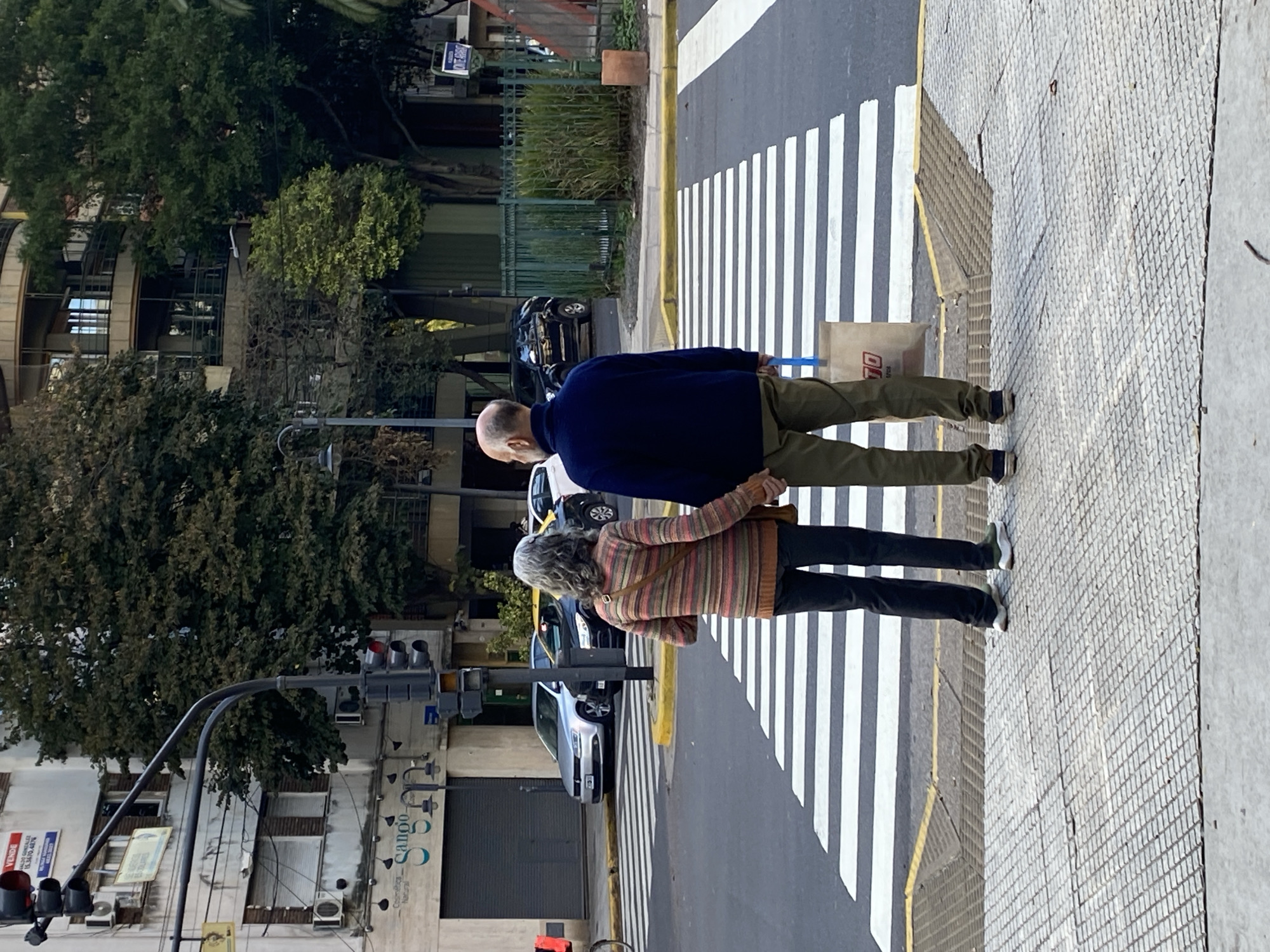 An older couple holds hands while crossing a pedestrian crossing on a street in Buenos Aires, Argentina. The man is wearing a blue sweater and tan pants, while the woman is wearing a striped sweater and dark pants. The street is lined with buildings, trees, and parked cars. A traffic light is seen in the foreground. The pavement is tiled with a patterned texture. There is a paper bag with what looks like blue handle near the couple.