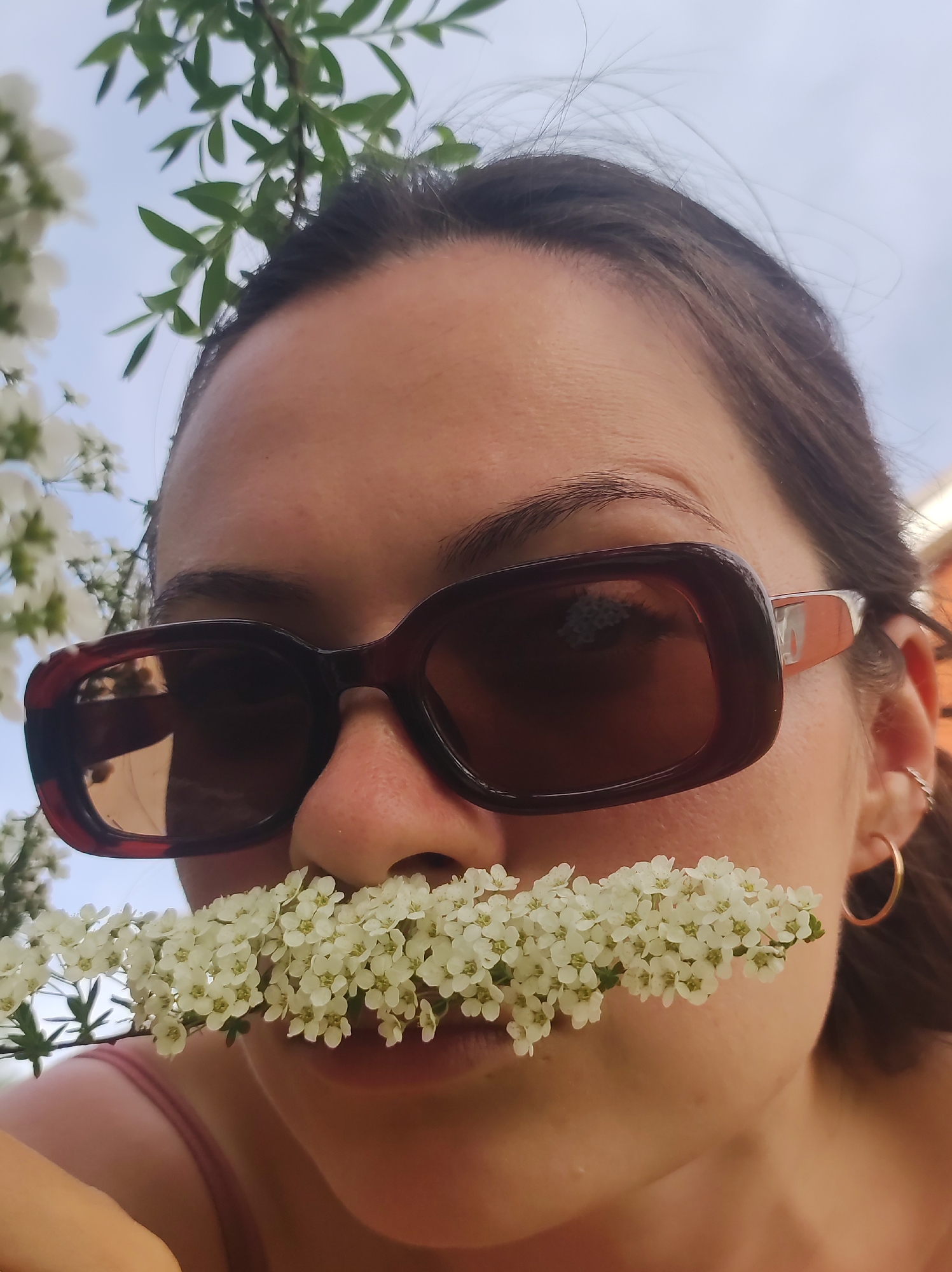 A person with dark hair and wearing brown sunglasses is close to the camera. A sprig of small white flowers is held horizontally between the nose and mouth. A gold hoop earring is visible on the right ear. Green leaves and sky are visible in the background. The location is not discernible from the visual information.