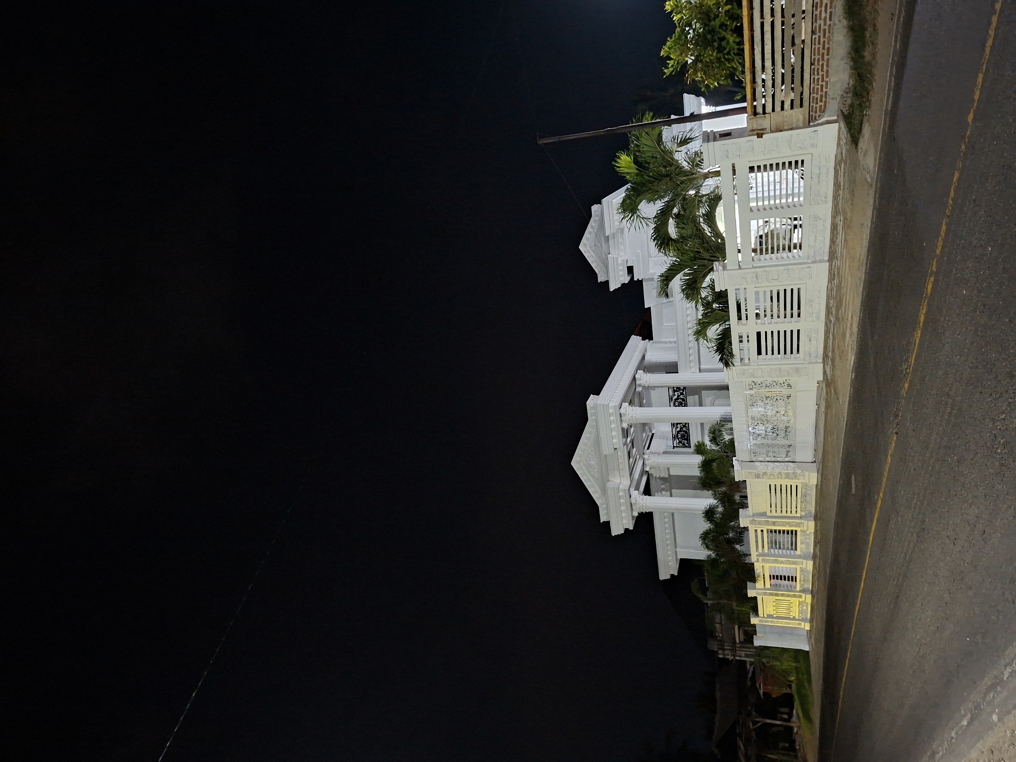 At night in Lancang Barat, Indonesia, a brightly illuminated white building stands adjacent to a paved road with a yellow center line. The building has multiple levels, with decorative lattice work and pillars. Palm trees grow near the building. A power line is visible against the dark sky.