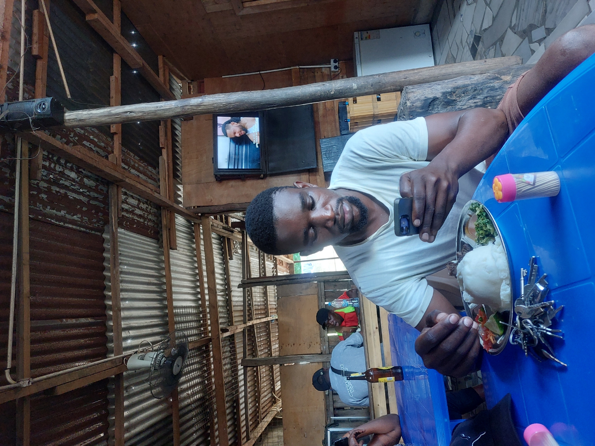 The image displays the interior of an informal eatery in Dar es-Salaam, Tanzania. A Black male, wearing a light-colored t-shirt and brown shorts, is seated at a bright blue plastic table in the foreground, holding a dark smartphone in both hands and looking down at its screen. A metal plate with a meal consisting of a large mound of white food (likely ugali), a portion of meat, and a small side salad is positioned directly in front of him. To his left on the table are a large cluster of keys, a black baseball cap, and a pink and orange plastic toothpick dispenser. A brown glass bottle is also present on the table, partially obscured by an arm of an adjacent, unseen person.
In the mid-ground, two other males are visible behind wooden partitions. One male, wearing a grey t-shirt with "SERENGETI" visible on the back and a black baseball cap, has his back to the camera. Another male, wearing a red shirt and a black baseball cap, faces partially away, with a clear plastic water bottle on the table in front of him.
The background wall is composed of light brown wooden panels. A flat-screen television is mounted on this wall, displaying a bearded male figure with partial subtitles visible. Below this, a second, darker screen or unit is present. A robust, unfinished wooden post extends from floor to ceiling, with a black speaker mounted on it near the top. To the far right, a white upright refrigerator is visible. The floor is covered with irregularly shaped, light-colored tiles.
The ceiling consists of exposed wooden beams supporting corrugated metal sheets, some of which show signs of rust. A white oscillating fan is suspended from the wooden structure. White electrical conduits are also visible running along the ceiling.