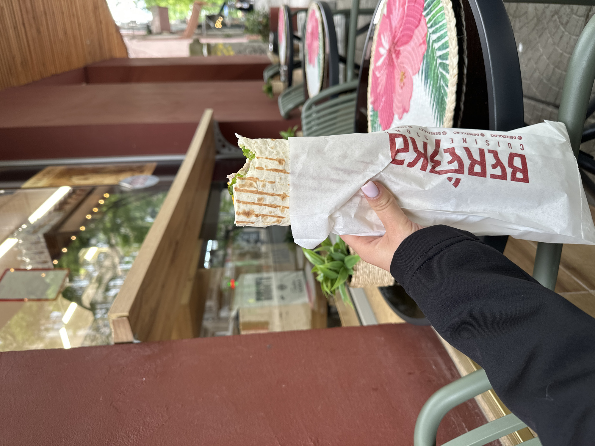 A person wearing a black long-sleeved shirt is holding a Berezka Cuisine sandwich wrapped in a white paper bag. The sandwich is partly eaten and shows fillings like lettuce and sauce. The Berezka Cuisine logo is printed in red on the bag, with the website berezka.bg and address berezka.ro also visible. The background features outdoor cafe-style seating with chairs that have flower designs and a wooden structure with a red-brown painted facade. Reflective glass can be seen behind the wooden structure. The location is likely in the city of Sofia, Bulgaria, based on the restaurant's branding, though specific landmarks are not visible in the image.