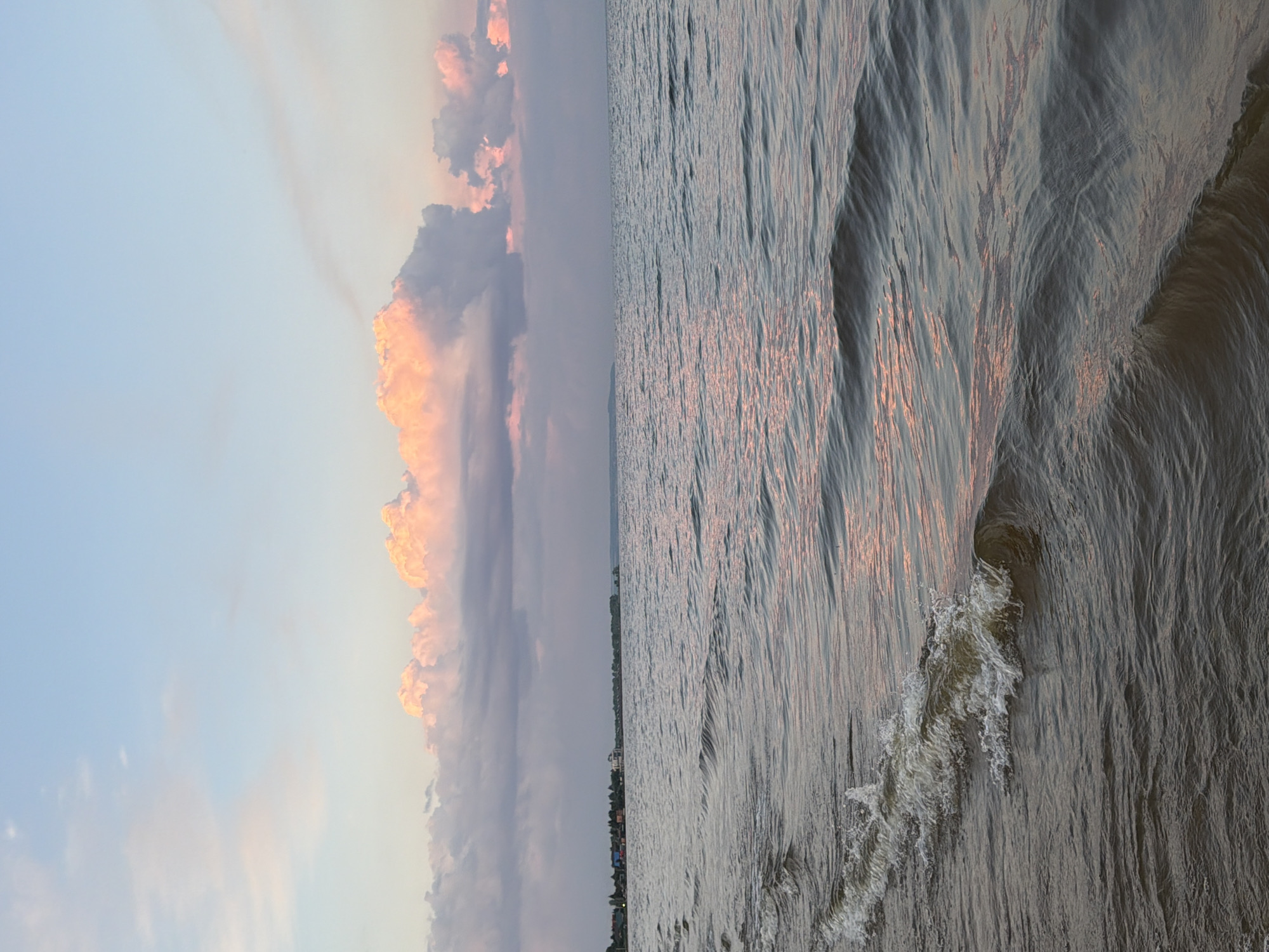 A wide shot captures a serene seascape with a horizon line dividing the water and the sky. The water, in the foreground and midground, is a deep, muted blue-grey, with subtle ripples and gentle waves breaking near the left edge. The surface of the water reflects the soft hues of the sky. In the distance, a faint shoreline is visible, with a line of buildings and trees. The sky above is a soft gradient of pale blue, transitioning to hues of pink and orange near the horizon, where large, fluffy clouds are illuminated with warm, rosy light. The overall impression is one of tranquility and natural beauty. This scene is located in Laishevo, Russia.