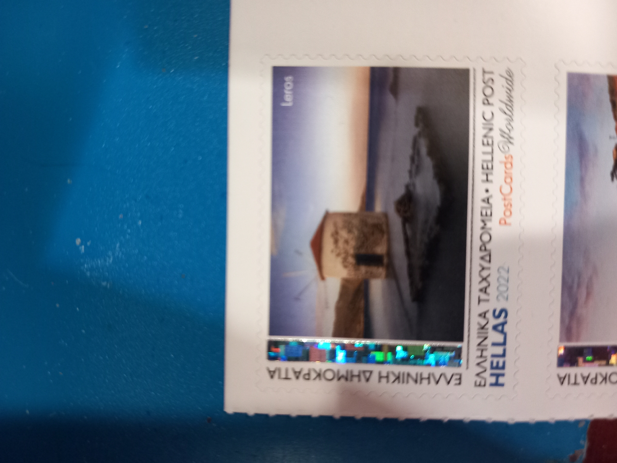 The image shows a close-up of a postage stamp with a scenic depiction. The stamp is part of a larger sheet, indicated by perforated edges and text. The visible portion of the stamp features a round, stone building with a red roof, situated on a rocky coastline. The water in the background appears calm, and the sky shows a sunset or sunrise with soft, warm hues. The text on the stamp includes "LEROS" in the upper right corner, and below the image, "ΕΛΛΗΝΙΚΑ ΤΑΧΥΔΡΟΜΕΙΑ HELLENIC POST HELLAS 2022 PostCards Worldwide." To the left of the stamp image, vertical text reads "ΕΛΛΗΝΙΚΗ ΔΗΜΟΚΡΑΤΙΑ," and below that, partially obscured, "ΟΚΡΑΤΙΑ." The background behind the stamp sheet is a textured, blue surface, possibly a table or wall. The stamp itself has a holographic or iridescent strip along its left edge.