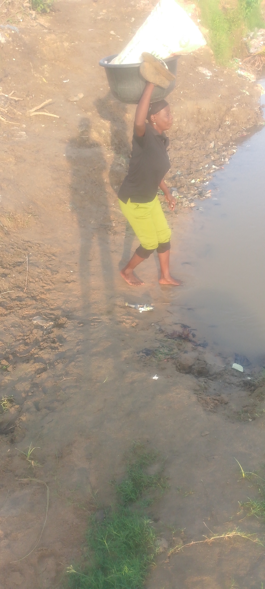 In Jalingo, Nigeria, a woman is captured in a vertical, full-body shot as she walks barefoot at the edge of a body of water. She is carrying a large, dark basin filled with a white substance on her head, balanced with her right hand. The woman is wearing a dark t-shirt and bright lime green shorts. Her shadow is cast on the muddy ground to her left. The environment appears to be a natural setting, with dry, sandy earth and some sparse vegetation near the water's edge. The water itself is murky, and there is visible debris, including plastic waste and other refuse, along the bank and floating in the water. The lighting suggests it is daytime, possibly late afternoon or early morning, with the sun casting long shadows.