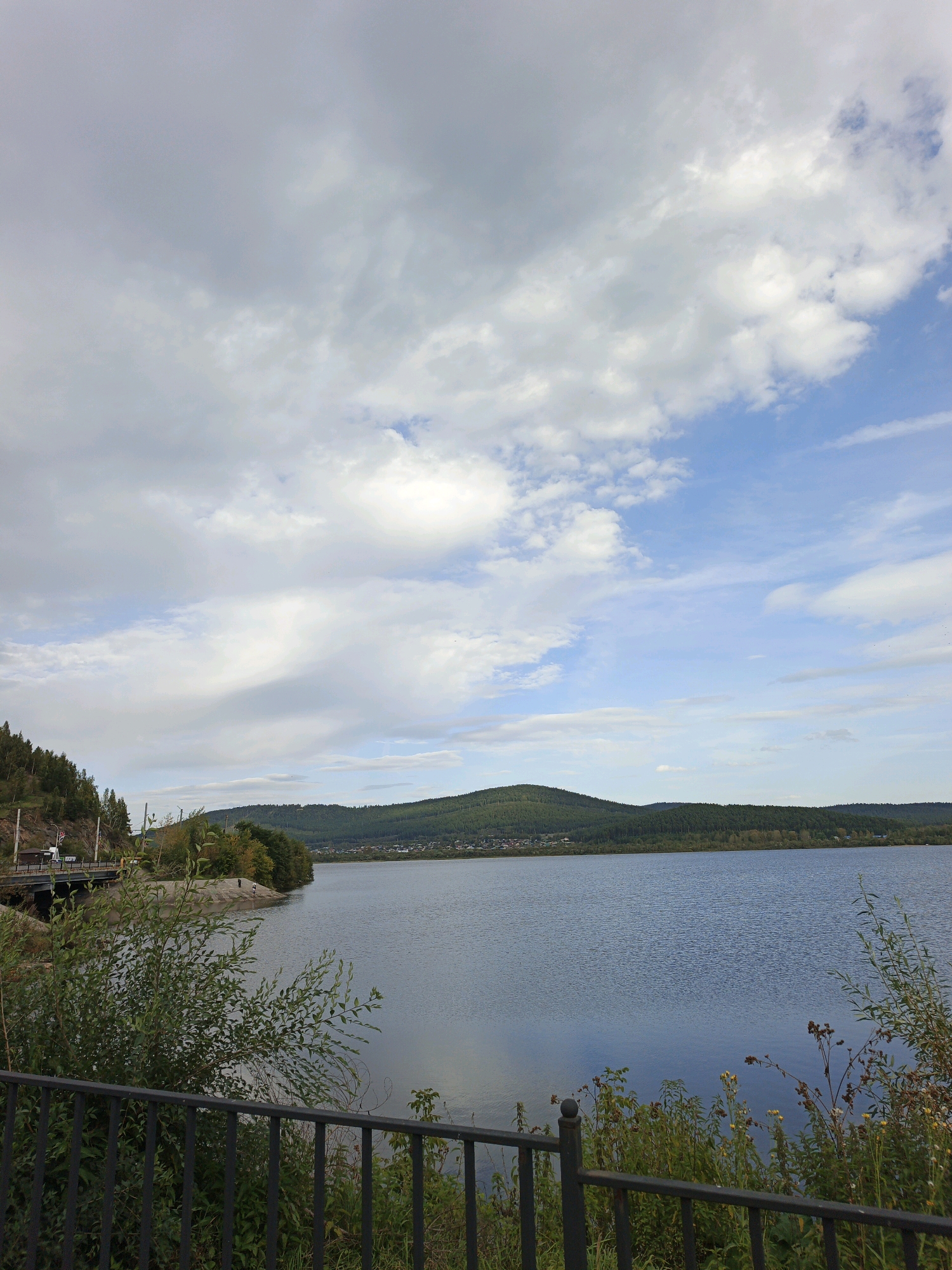 The image captures a serene landscape of a large body of water, likely a reservoir, on a cloudy day. In the foreground, a black metal fence runs horizontally across the bottom of the frame, with lush green foliage and some dry plants growing behind it. The water stretches out towards a distant, tree-covered hill range. The hills are densely forested, with a small settlement visible on their slopes. The sky is filled with dramatic, cumulus clouds, with patches of blue sky peeking through. On the left side of the image, a bridge can be seen crossing a section of land that leads to the water's edge. A few trees with autumn colors are scattered along the embankment. The scene appears to be located near Zlatoust, Russia, given the characteristic landscape of the Ural Mountains region. There are no visible people or specific actions taking place in the image, creating a peaceful and natural ambiance.