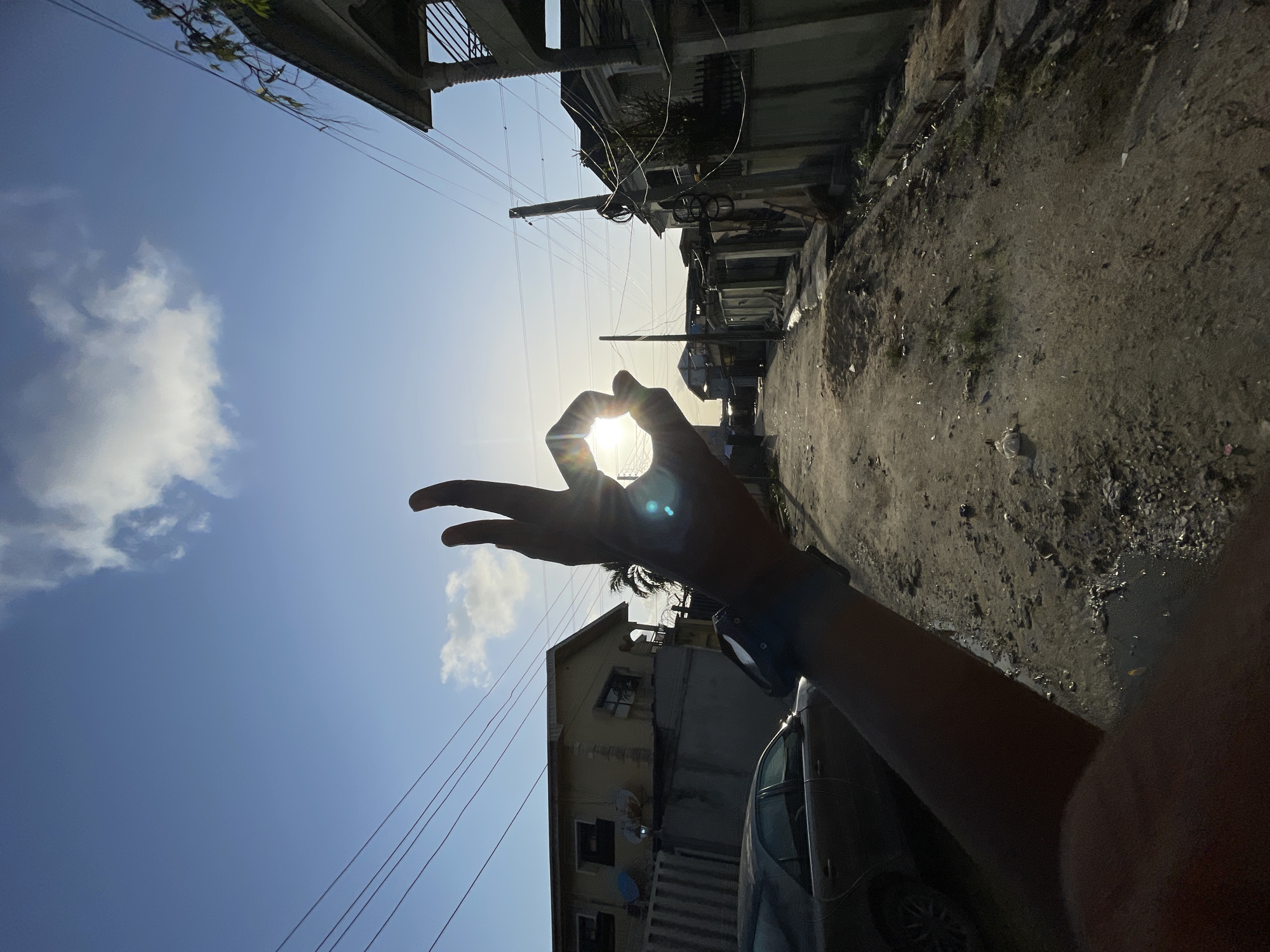 The image shows a street scene in Ado, Nigeria, oriented vertically with the camera pointed upwards at an angle. The sun is positioned centrally, partially obscured by a hand making an "OK" sign. A blue smartwatch is visible on the wrist. Buildings line the sides of the street, which appears unpaved and somewhat dilapidated. Overhead power lines crisscross the sky. A light-colored vehicle, possibly a car, is partially visible in the foreground. The sky is blue with some scattered clouds. The lighting indicates it is daytime.