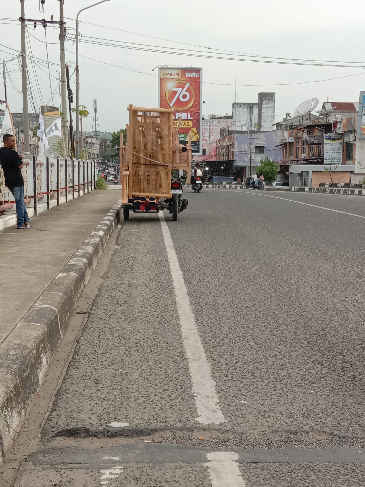 The image captures a street scene in Lhokseumawe, Indonesia, depicting a relatively wide asphalt road with a solid white lane marking. A pedestrian sidewalk bordered by a low, decorative white concrete fence runs along the left side of the road. A man in a black shirt and blue jeans stands on the sidewalk, leaning against the fence.
To the right of the man, partially on the sidewalk and partially on the road, is a motorcycle with a large, square bamboo frame attached to its rear. The frame is loaded with soft, red and black cushions or pillows. The motorcycle faces forward down the road.
In the background, buildings of varying heights and designs line the street, with a large billboard featuring the number "76" visible above the structures. Additional motorcycles and scooters are seen navigating the road in the distance. Overhead power lines and utility poles are present along the side of the road. The sky is overcast, suggesting a cloudy day.