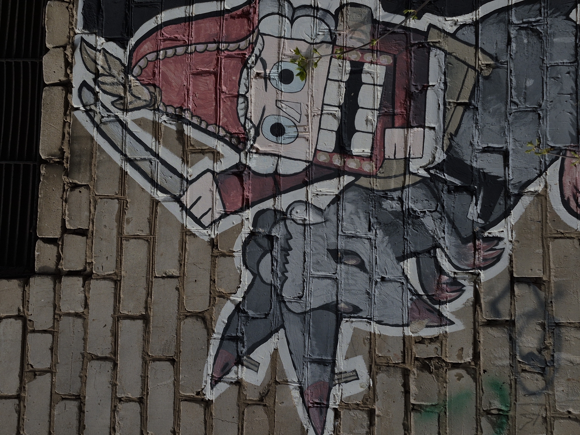 Graffiti-style cartoon imagery is painted on a brick wall. A stylized figure, primarily rendered in pink, red, black, white, and shades of gray, appears to be a surreal or abstract representation of a person. The upper portion features a large, open mouth with prominent teeth, stylized eyes, and other facial features. Below this is a grey body, somewhat anthropomorphic, with abstracted limbs and garments.
The brick wall serves as the canvas for the artwork, and its texture is visible through the paint. The graffiti is found on a building in the city of Chaikovsky, Russia. The presence of a small plant growing in a crack on the wall suggests the image has been present for some time. A black metal grate is partially visible on the left side.