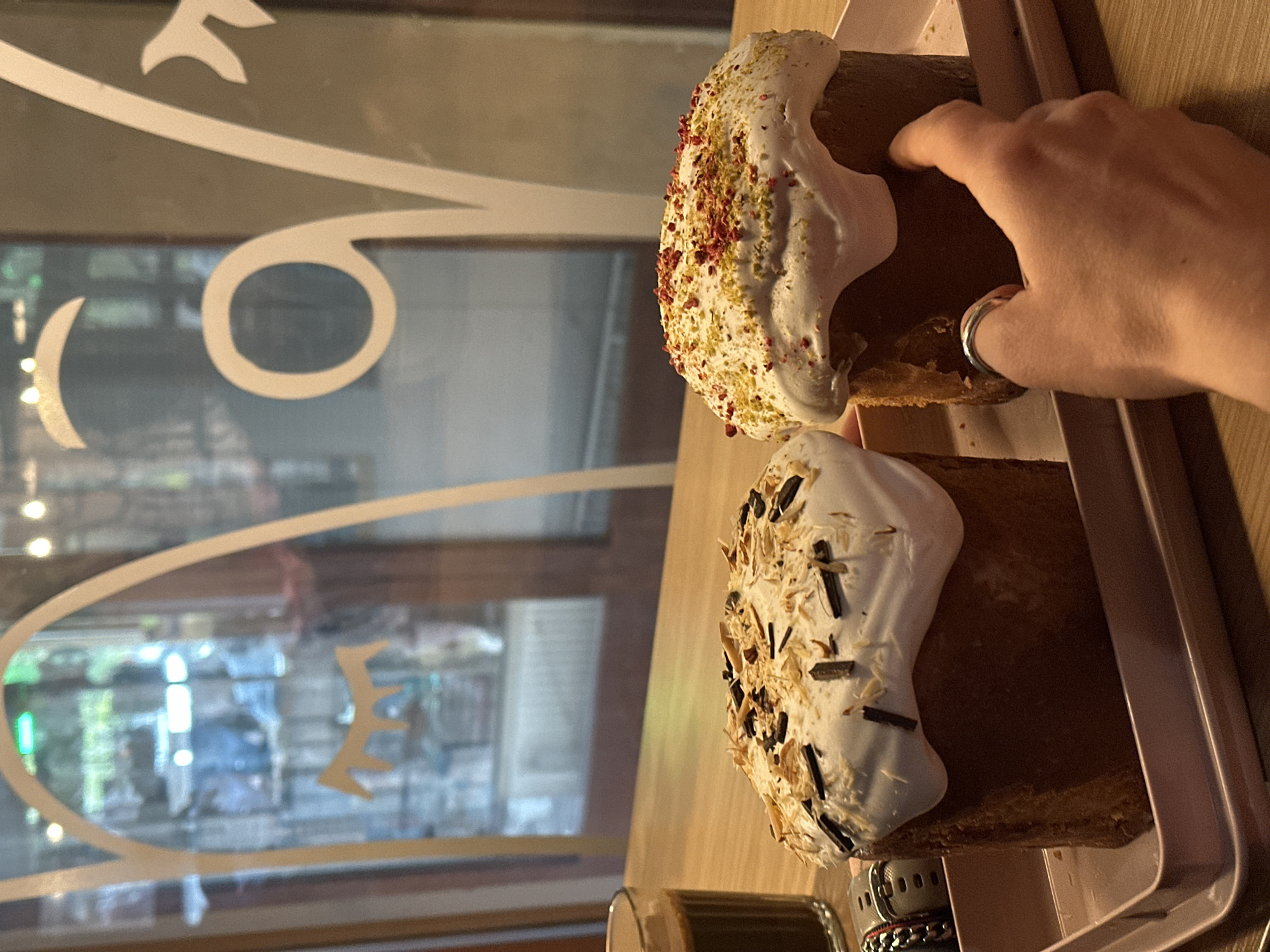 The photograph depicts a close-up view of two Easter cakes, also known as "kulich," placed on a pink tray. The cakes are decorated with white icing and various toppings, one featuring what appears to be pistachios and dried berries, while the other has shredded almonds and chocolate sticks. A person's hand, adorned with a ring, is visible near the top cake, possibly holding or reaching for it. In the blurred background, a window displays an out-of-focus street scene, including a logo or design on the glass. Based on the architecture visible, the location is likely a cafe or bakery in the city of Novi Sad, Serbia.