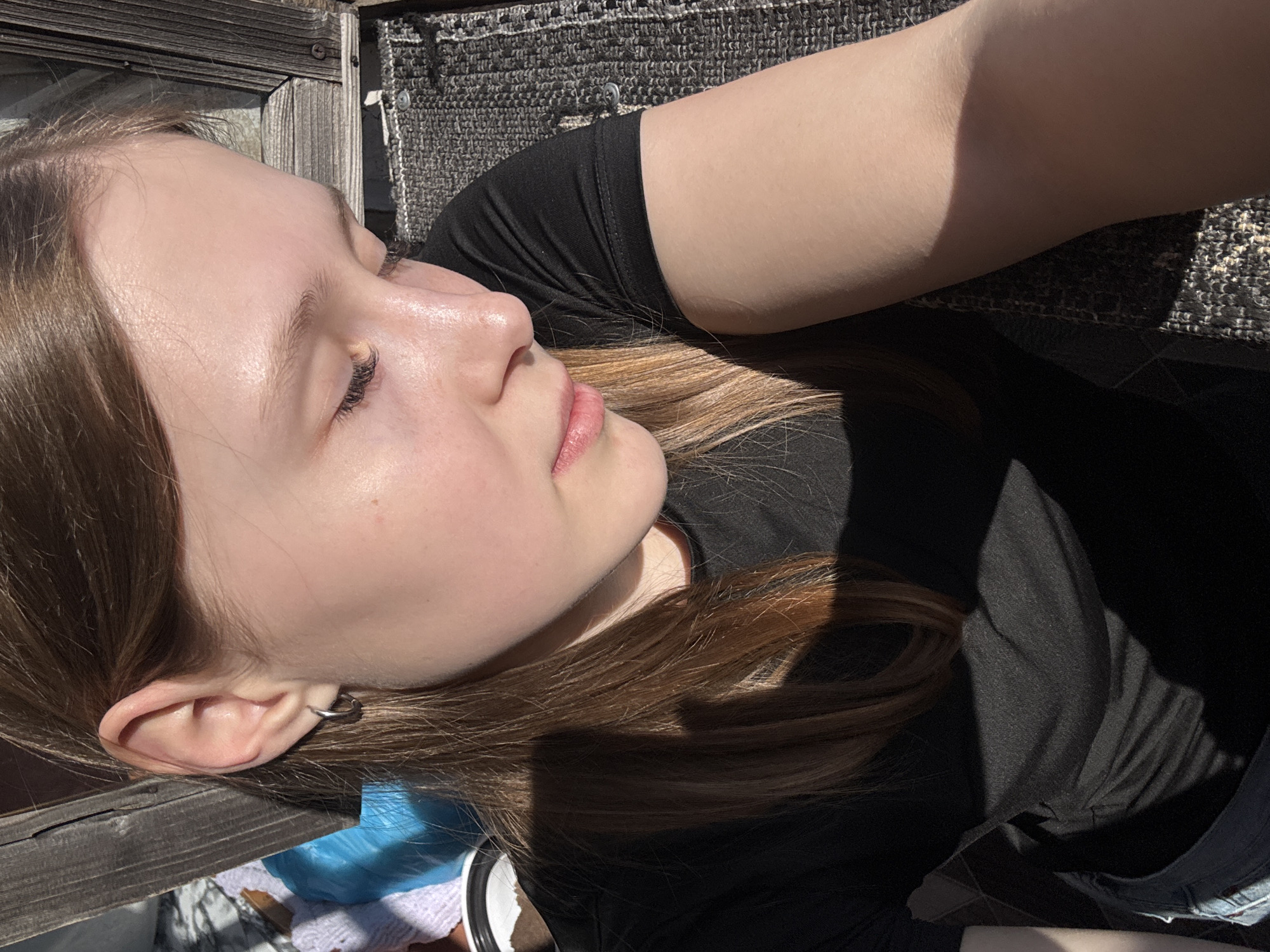 A fair-skinned young woman with light brown hair is lying down, presumably outdoors. She is wearing a black t-shirt. Her eyes are closed, and her face is turned towards the light. She has a hoop earring in her left ear. The background includes a woven mat or rug and what appears to be a wooden structure. The setting is unknown, and there is no indication that the picture was taken in the city of Chaikovsky, Russia.