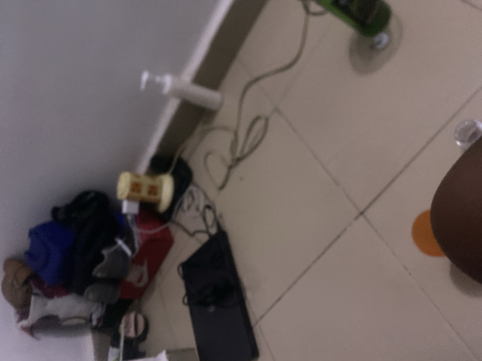 The image depicts a cluttered corner of a room, likely a bedroom or storage area, with items scattered on the tiled floor and against the wall. In the background, a pile of clothing and bags is visible, including a blue item that appears to be a duffel bag, a red Nike shoe box, and other miscellaneous items. To the right of the clothing pile, a yellow object with a patterned surface sits on a white base, possibly a lamp or air freshener. Further to the right, a white pump bottle, likely soap or lotion, stands on the floor. A black laptop is placed on the floor in the foreground, with cables extending from it. A green bottle, possibly a cleaning product or beverage, is also visible on the floor in the lower right section of the image, resting on a coaster. The floor is tiled with a beige or light brown color, and a dark shadow suggests the presence of an unseen object or person. An orange circular object and part of a dark brown object, likely a person's leg, are in the bottom center of the frame. The overall lighting is dim, and the image is somewhat blurry, suggesting it was taken with a low-quality camera or in low light conditions. The specific location in Howayi, Nigeria, cannot be determined from the visual information provided in the image.