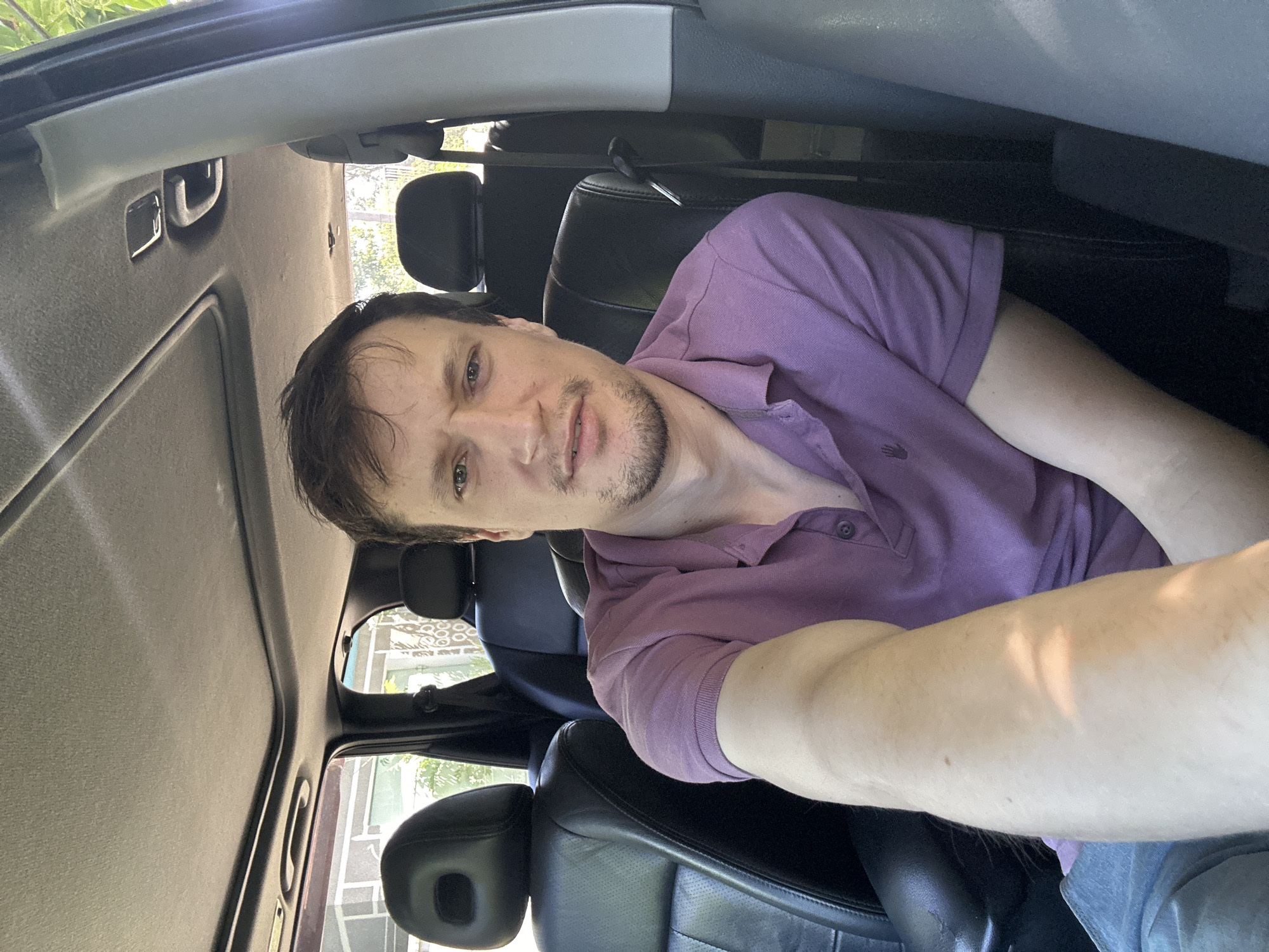 A man is captured in a selfie, seated in the passenger seat of a car. He is wearing a purple polo shirt and blue jeans. The interior of the car is visible, including the black leather seats, the headrests, the sunroof, and a portion of the dashboard and pillar. The man is looking directly at the camera with a neutral expression. The scene appears to be during the daytime, with sunlight filtering into the car. The location is identified as Makiv, Ukraine.