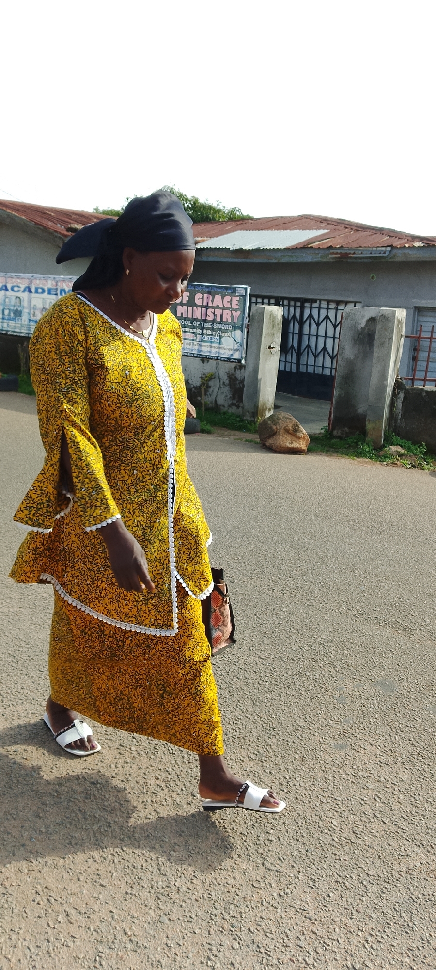 An adult African woman walks on an asphalt road in front of a building. She is wearing a yellow dress with a black floral pattern, a black headscarf, and white sandals. She is carrying a patterned bag. The building behind her has a metal gate and a sign that reads "OF GRACE MINISTRY". There is a large rock on the ground to the right of the building entrance. The sky is overcast and white. The overall lighting suggests it is daytime.