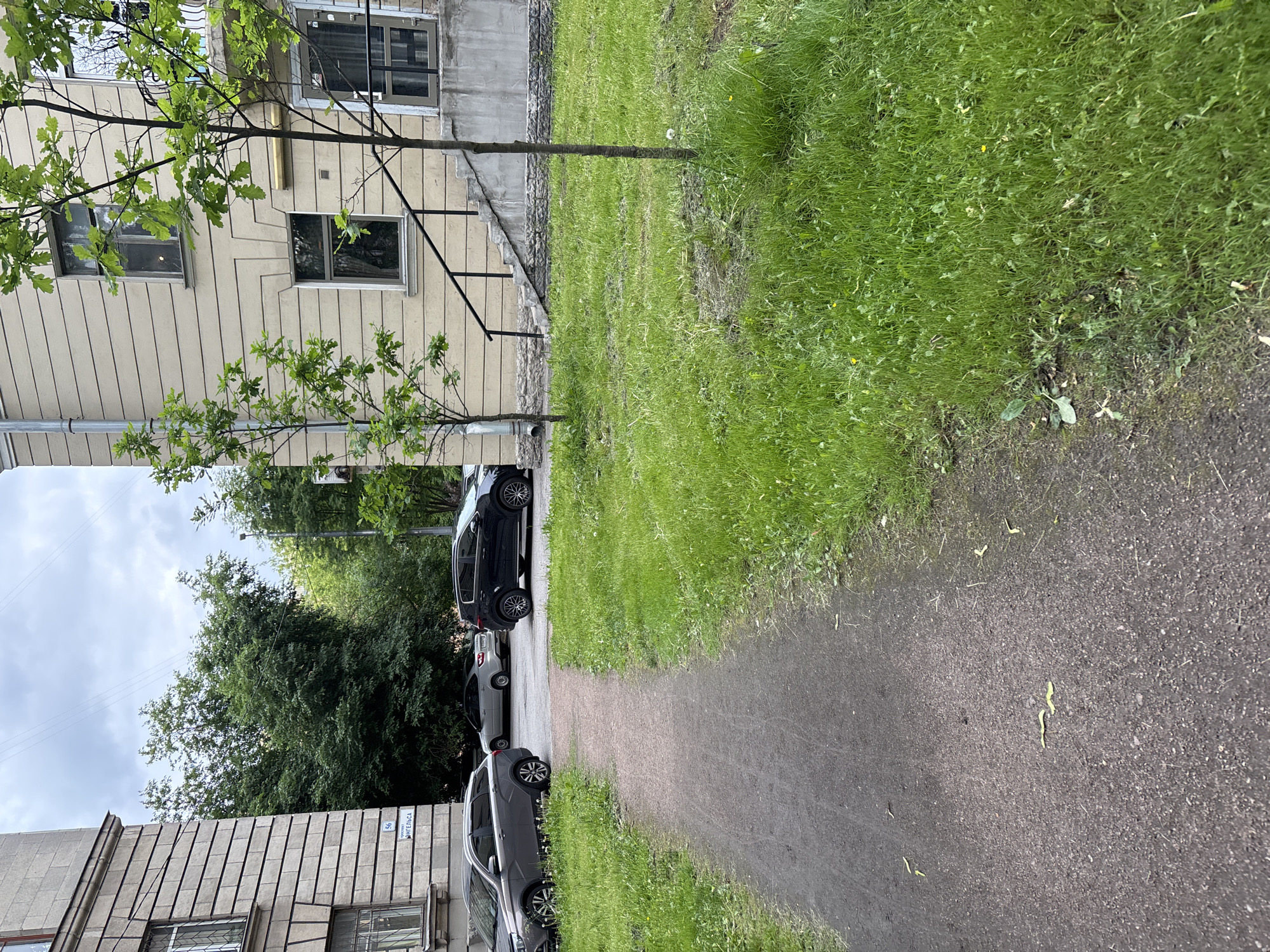 The image shows a view of a residential street in Saint Petersburg, Russia. The scene is dominated by a building with a light-colored facade, featuring several windows and a prominent staircase leading to an entrance. In front of the building, a patch of mown grass is visible, with a pathway cutting through it. Several parked cars are present on the street, including a dark SUV and a silver hatchback. Trees with green foliage are scattered throughout the area, adding to the urban greenery. The sky is overcast, suggesting a cloudy day. The overall impression is that of a typical urban residential setting in Saint Petersburg, with a focus on the architectural features of the building and the surrounding environment.
