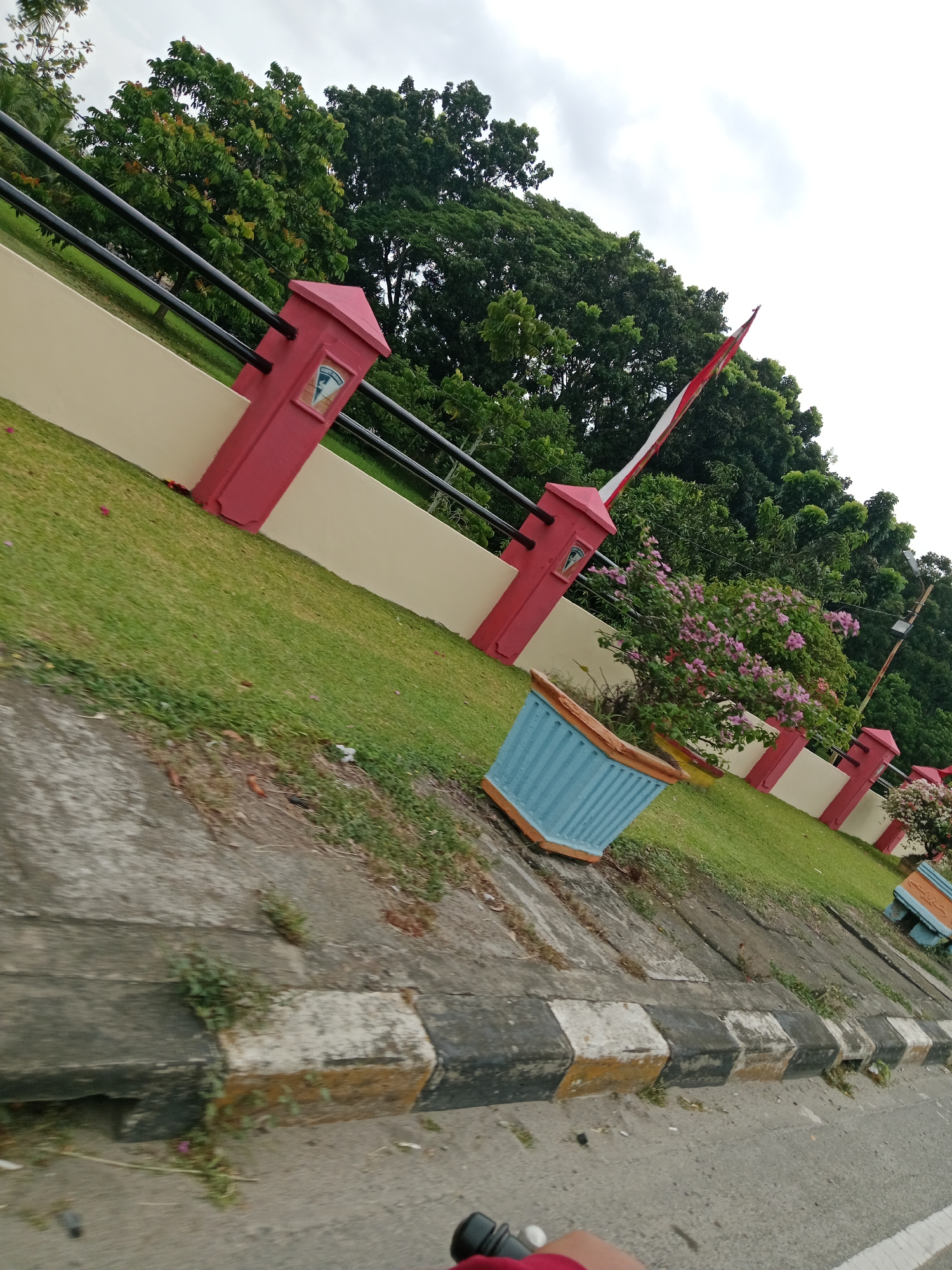 The image depicts a street-level view of a red and cream-colored wall with a black metal fence. The wall is punctuated by red pillars topped with what appears to be a coat of arms. A red and white striped barrier arm is visible extending from one of the pillars. Lush green grass lines the area behind the wall, with a vibrant pink flowering bush in a blue and brown planter. Further down, another planter with pink flowers is visible. The foreground shows a concrete sidewalk with alternating black, white, and yellow markings. The perspective suggests the photo was taken from a moving vehicle, possibly a motorcycle, as indicated by the blurry foreground and the partial view of a red garment and black handlebars in the bottom right corner. The sky is overcast. The scene is likely a public space or the entrance to a facility. Based on visual cues from the context of "City of Binjai, Indonesia," this could be an entrance to a government building, park, or a public facility within the city.
