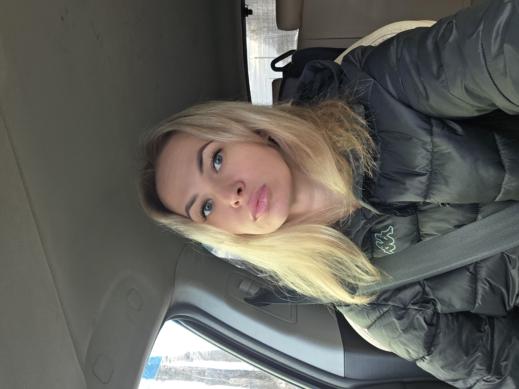 A young woman with blonde hair and green eyes is seated in a car, wearing a black puffer jacket with a seatbelt fastened. The interior of the car is visible, including the door panel and headliner. Outside the window, it appears to be a winter scene with snow and bare trees. The location is not explicitly discernible as Chaikovsky, Russia, based on the provided image.