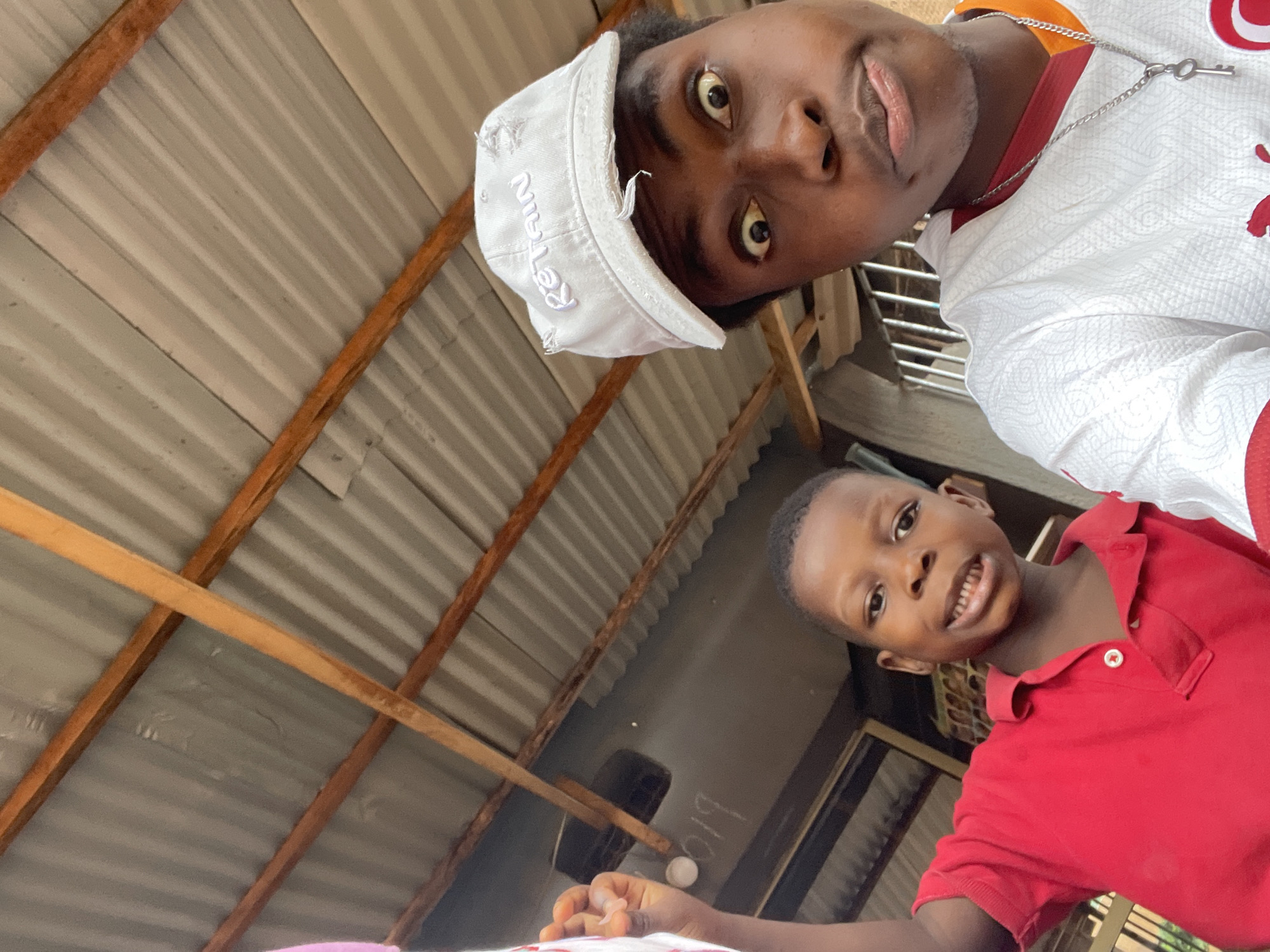 Two individuals, a youth and a child, are depicted in an image taken from a low angle. The youth is wearing a white cap with distressed fabric, a white shirt with red trim, and a silver necklace with a key pendant. The child is wearing a red polo shirt. The background features a corrugated metal roof with wooden supports and partial view of a dark wall with white lettering. The location is identified as Onitsha, Nigeria.