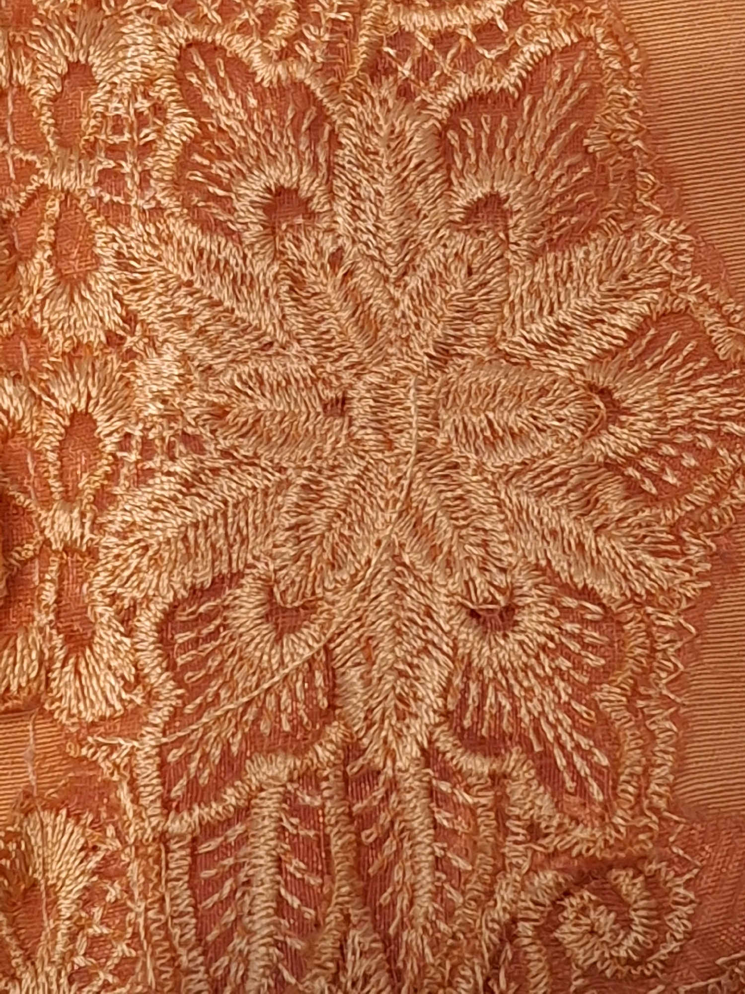 The image displays a close-up view of an orange embroidered fabric. The embroidery features a floral or mandala-like pattern with intricate details and a textured, almost feathery appearance. The color is a warm, earthy orange. There are no people, actions, or interactions visible in the image. The environment is not discernible beyond the fabric itself. This image does not contain any elements that can be used to determine its location in Lumajang, Indonesia.