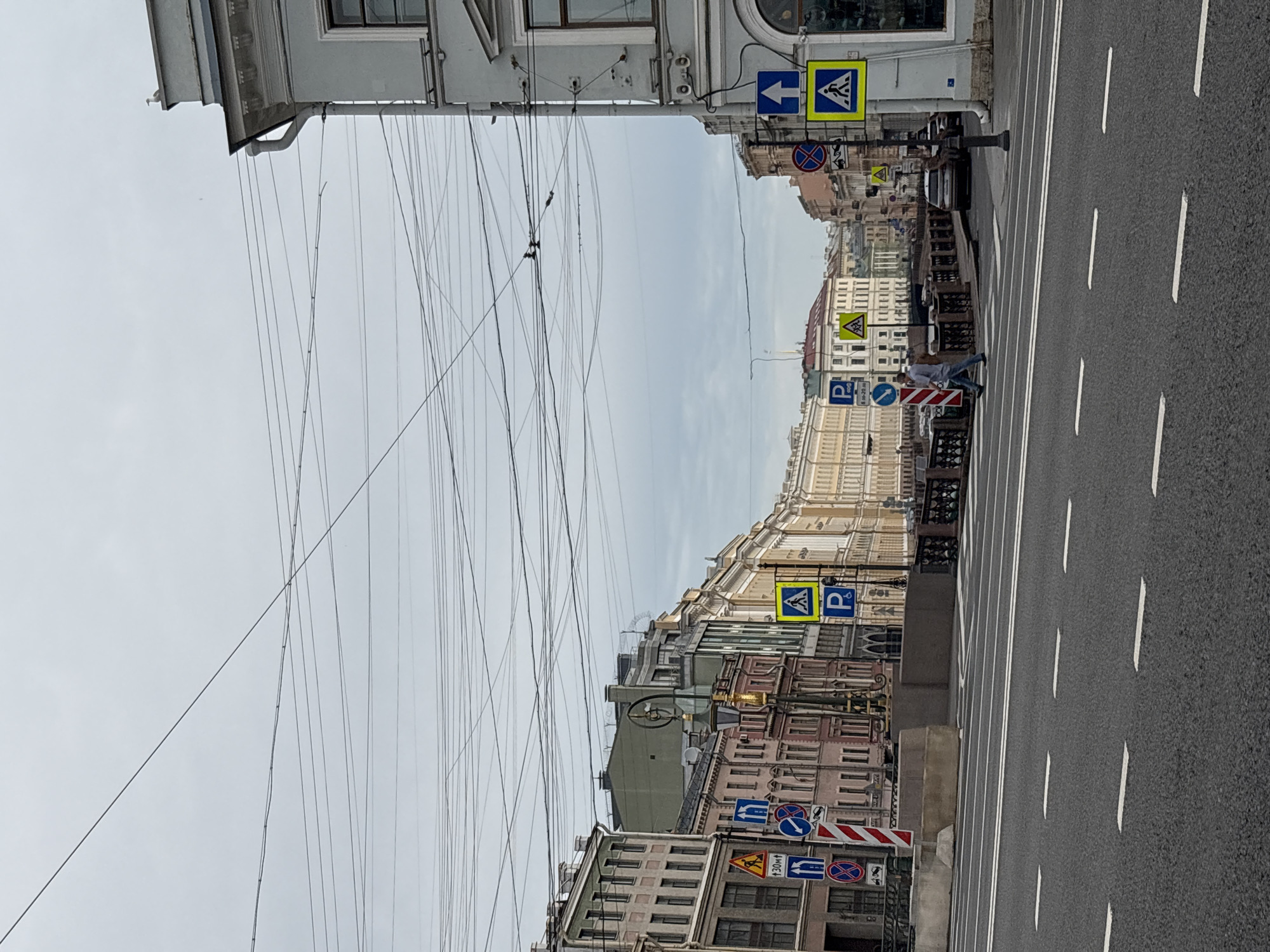 The image depicts a street scene in Saint Petersburg, Russia, likely on a cloudy day. In the foreground, a wide asphalt road with white lane markings is visible. A pedestrian crossing is marked with white lines. In the middle ground, a bridge spans a canal, with buildings lining both sides of the waterway. The buildings are diverse in style and color, with some featuring pastel hues and others more muted tones. Traffic signs, including pedestrian crossing signs, directional arrows, and parking signs, are prominent along the street. A person is walking across the pedestrian crossing. In the background, more buildings extend into the distance, and a complex network of overhead wires crisscrosses the sky. The overall atmosphere is urban and somewhat subdued due to the overcast weather.