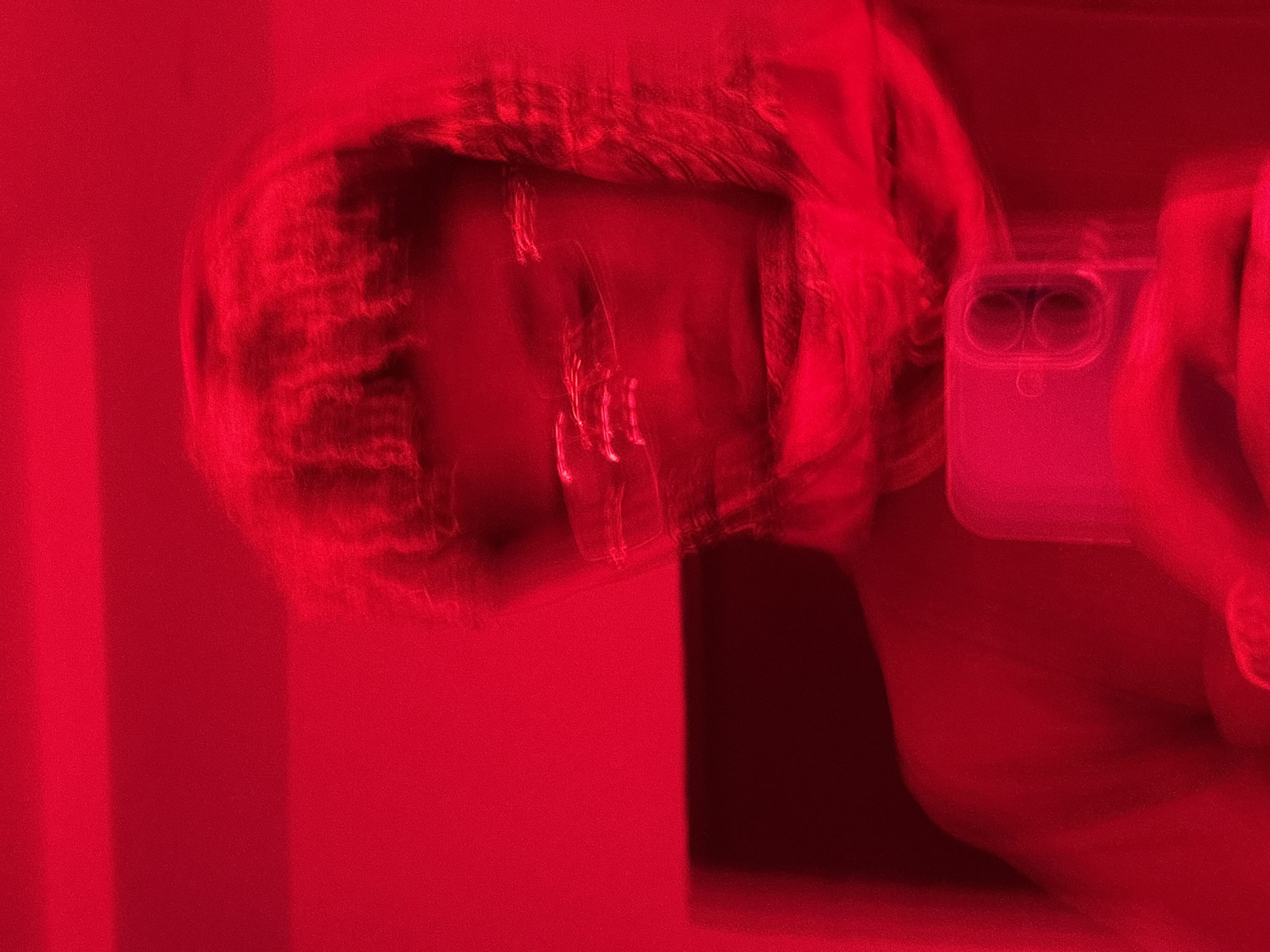 The image depicts a single individual in what appears to be an indoor setting in Igboefon, Nigeria, under intense red lighting conditions. The scene is significantly blurred, indicating motion or a long exposure. The individual, of indeterminate gender, is positioned centrally, facing forward, and holding a smartphone in their right hand (viewer's left). The smartphone, enclosed in a clear case, features a dual-lens camera system vertically aligned on its rear. The person wears clear-framed glasses, with reflections visible on the lenses. Their hair appears textured or styled in short dreadlocks/braids, blurred around the head. A dark garment, possibly a shirt or jacket, covers the torso, and a fabric, possibly a scarf, is draped around the neck and shoulder area. The background is a uniform red, with a faint horizontal dark band discernible in the upper section, possibly a shelf or part of a wall fixture. No other discernible objects or individuals are present.