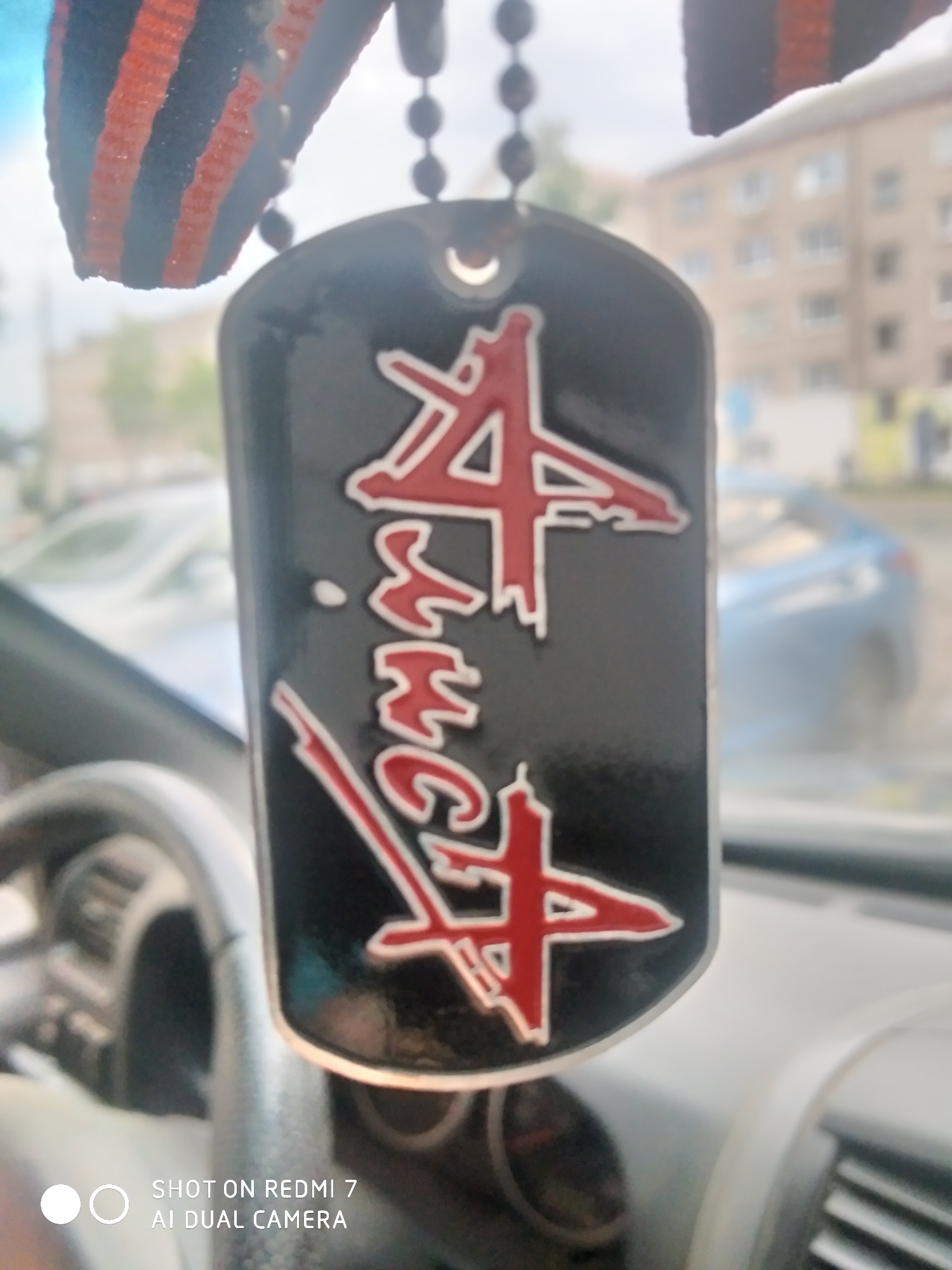 A close-up photograph shows a black dog tag with red calligraphy hanging from the rearview mirror inside a vehicle, likely a car. The tag is attached to a beaded chain. Above the tag, a ribbon with orange and black stripes is partially visible. In the background, seen through the windshield, is a blurry urban landscape including a residential building. The scene appears to be in Chaikovsky, Russia, though this can't be stated with certainty from the image alone. The bottom left corner of the image contains a watermark indicating the photo was "SHOT ON REDMI 7 AI DUAL CAMERA".