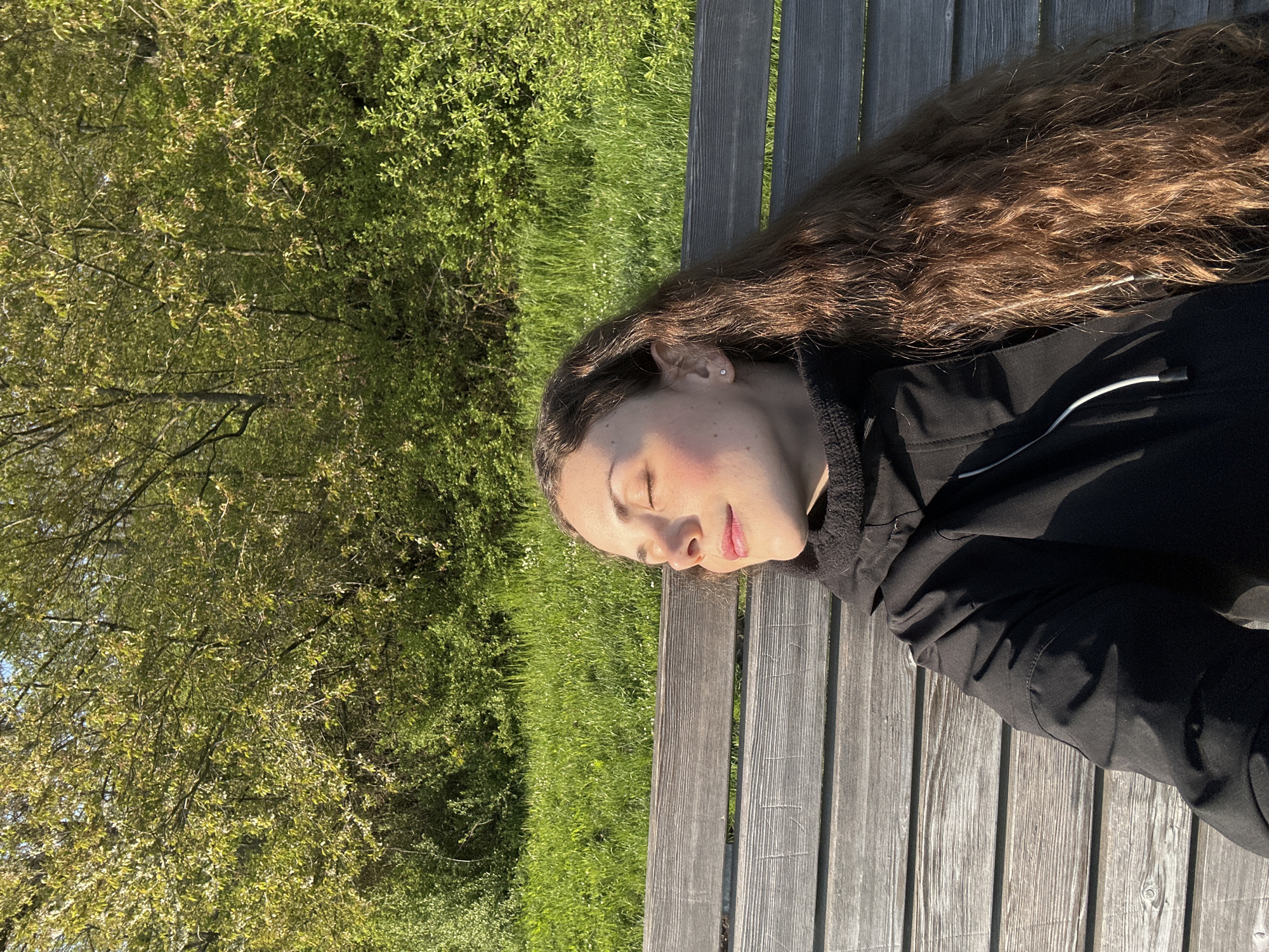 A young woman lies on a weathered wooden bench outdoors. Her eyes are closed, and she appears relaxed, possibly sleeping. She is wearing a black jacket with a stand-up collar, and her long, wavy brown hair spreads across the bench. The bench is made of parallel wooden planks with visible grain. To the left of the bench, there is a patch of green grass and a tree with dense foliage. The location appears to be a park or garden.