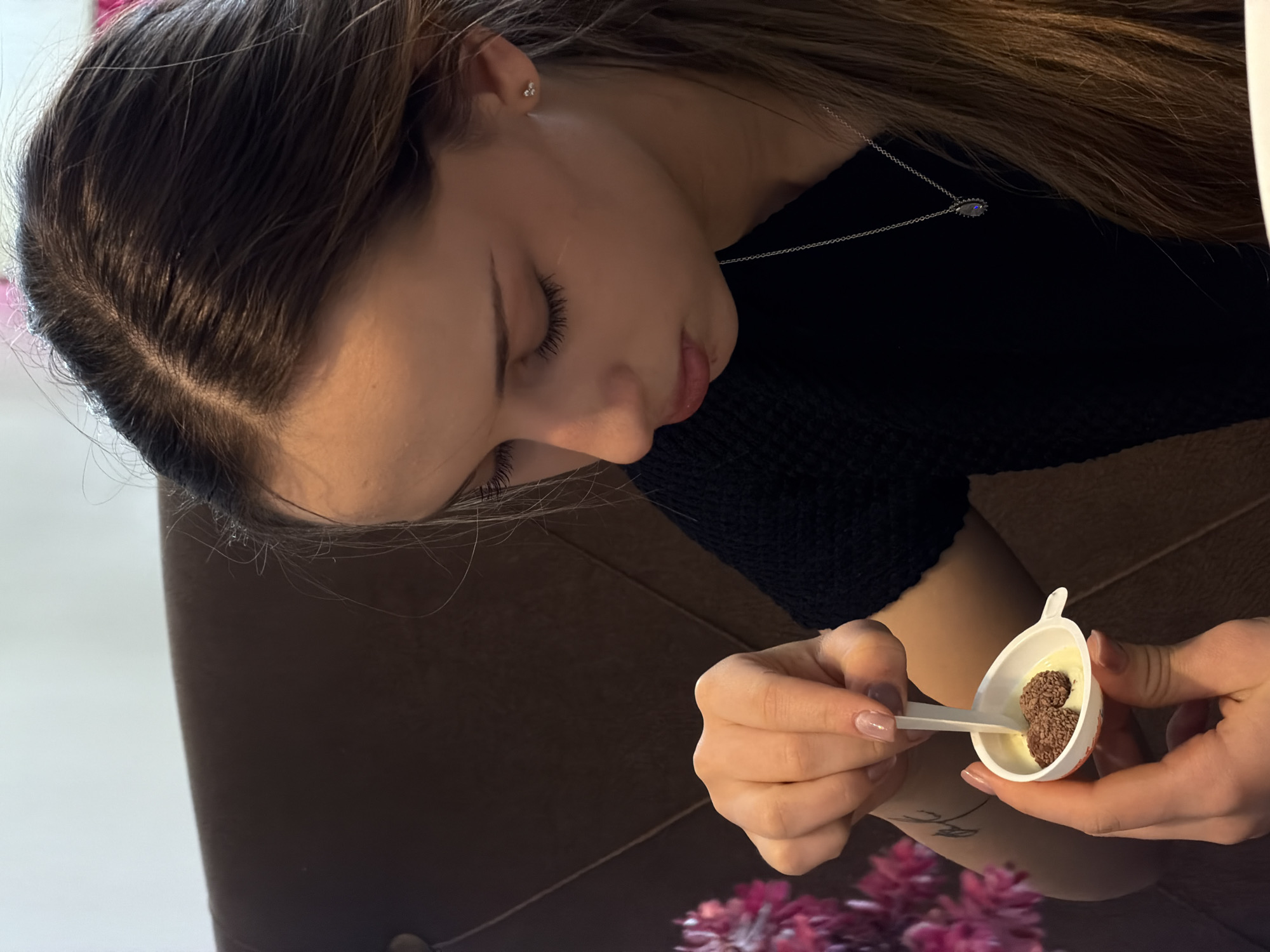 A woman is shown in a medium shot, her face directed towards a small, white cup held in her hands. She is using a white spoon to interact with the contents of the cup, which appear to be a light-colored creamy substance and two dark brown, spherical objects. The woman is wearing a black, textured shirt and a delicate silver necklace with a circular pendant. Her hair is long, straight, and brown, parted to the side. Her fingernails are manicured with a clear, glossy finish. A section of a brown leather couch is visible in the background. In the lower part of the image are pink flowers. The location is not discernible as Sofia, Bulgaria, from the visual elements present.