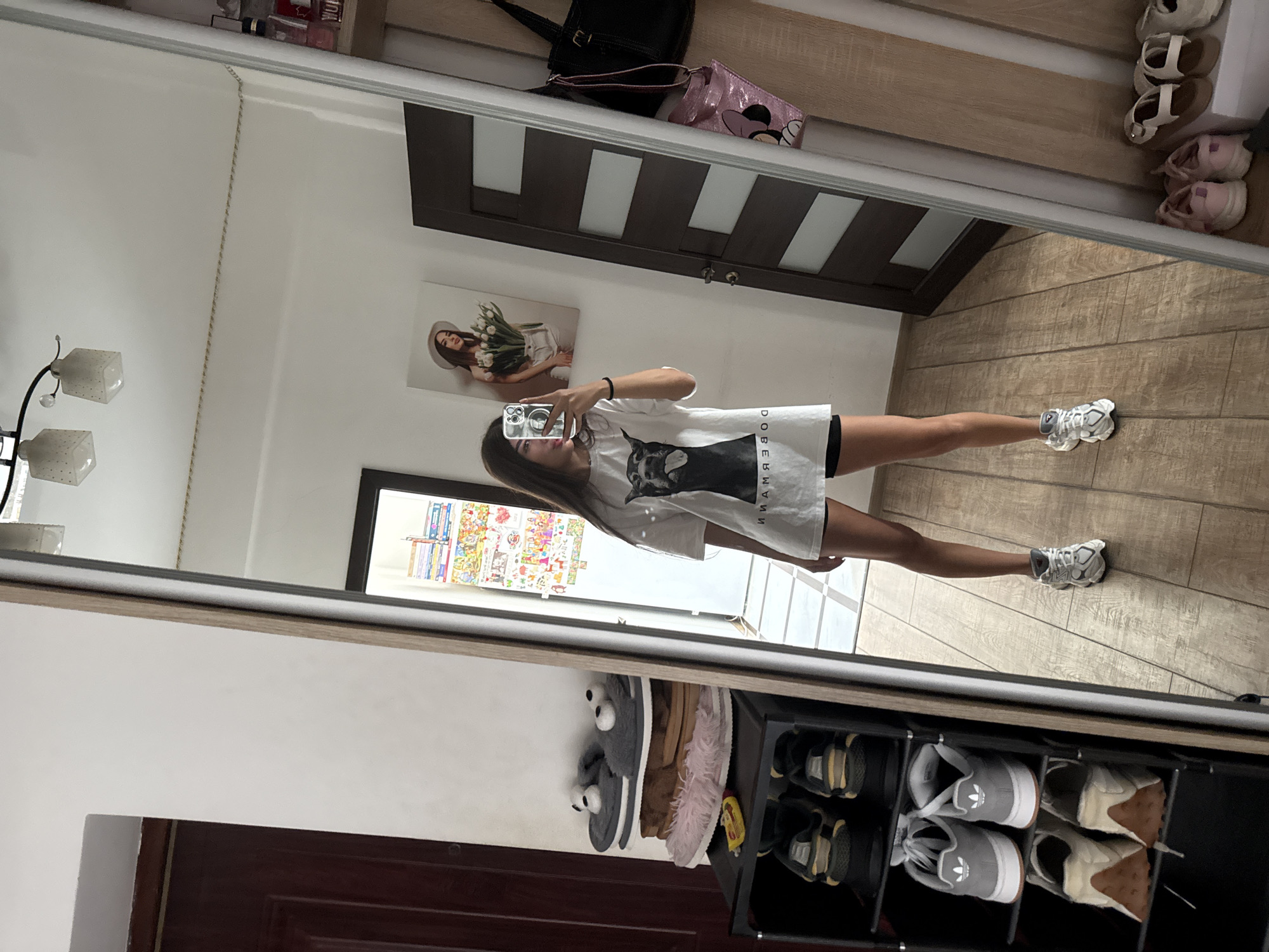 A young woman with long, dark hair is taking a mirror selfie in what appears to be a residential hallway or entryway. She is wearing a white t-shirt with a graphic print of a dog and black shorts. She has on white athletic shoes. The hallway has light-colored walls and wood-patterned flooring. To the left of the woman is a tall, narrow shoe rack filled with various pairs of footwear. Above the shoe rack, on the wall, are several pairs of fuzzy slippers, one gray pair with googly eyes resembling a bear. To the woman's right, there's a dark wood door with frosted glass panels and a large mirror reflecting the scene. On the wall near the door, a framed picture of a woman holding flowers is visible. Further to the right, the mirror reveals more of the hallway, including a shelf with some bottles and bags hanging on the wall. The overall impression is a casual, indoor setting. The presence of a shoe rack and the general decor suggest this could be an apartment or a house in Chișinău, Moldova.