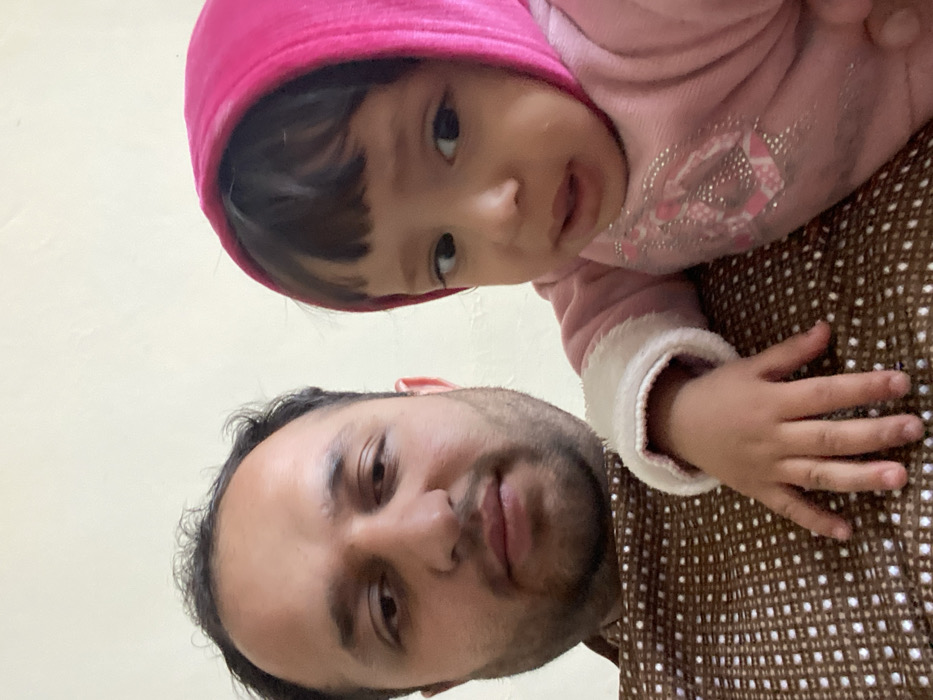 The image displays an adult male and a young child in a close-up indoor shot, originating from Kandahar, Afghanistan. The adult male, positioned to the left and slightly behind, possesses dark hair and a short beard. He wears a brown collared shirt with a repetitive white geometric pattern and faces directly towards the viewer. The child, situated to the right and in front of the adult, appears to be a toddler. This child wears a pink hooded garment featuring white trim on the sleeves and silver-colored embellishments on the left side of the chest/sleeve area. Dark bangs are visible beneath the hood. The child's left hand rests on the adult male's chest, and the child's gaze is directed towards the viewer with a slightly open mouth. The background is a plain, light-colored wall.