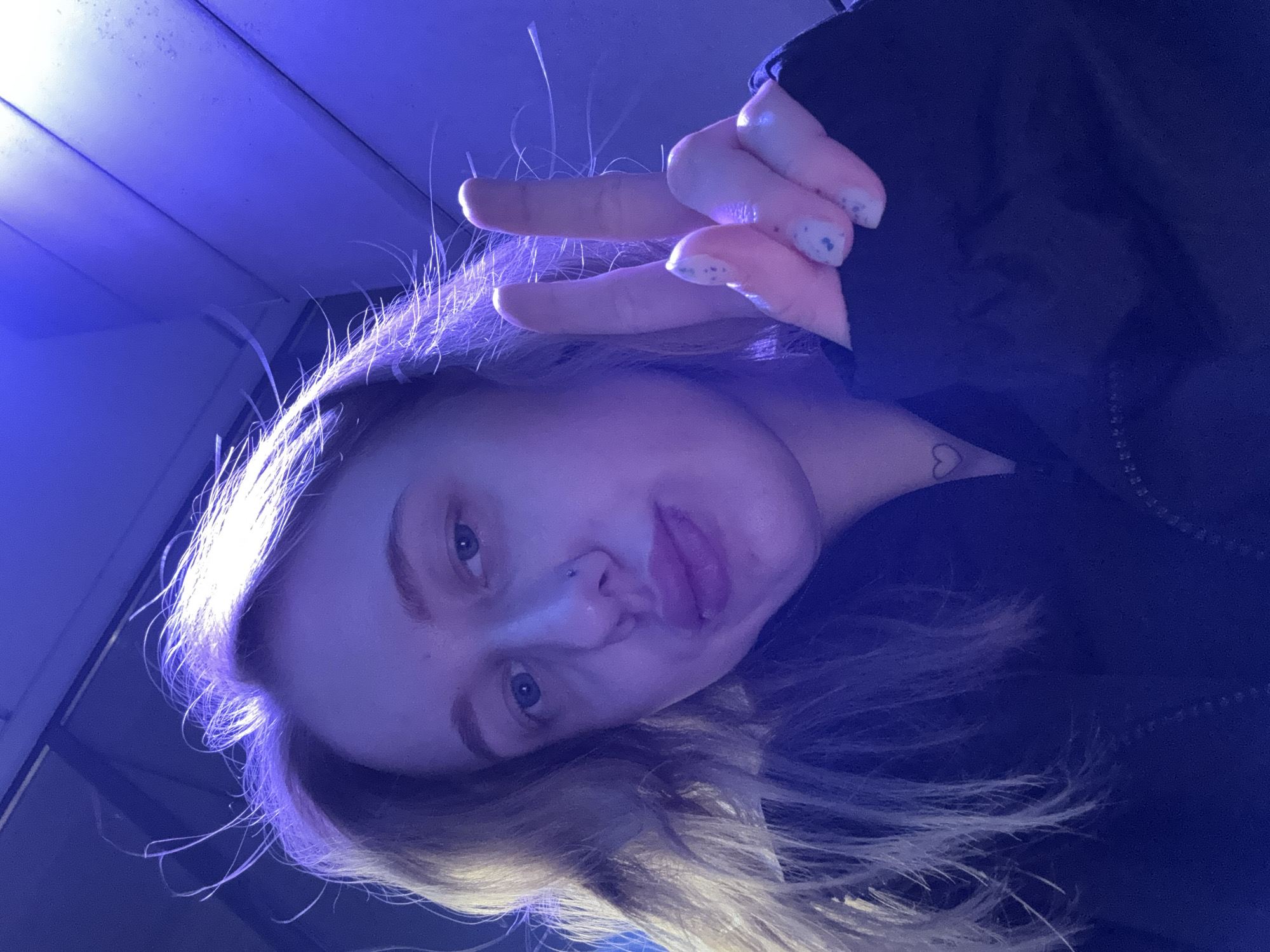 A selfie of a young white-skinned person with blonde hair. The person is holding up a hand, making a peace sign, with light blue nail polish. They have blue eyes, a nose piercing on the left side, and are sticking their tongue out slightly. A small heart tattoo is visible on their neck. The background appears to be indoors, possibly a room with blue lighting and white walls. It is impossible to verify a specific city, such as Ol'ginskaya, Russia, or any location without additional context or visual cues in the image.