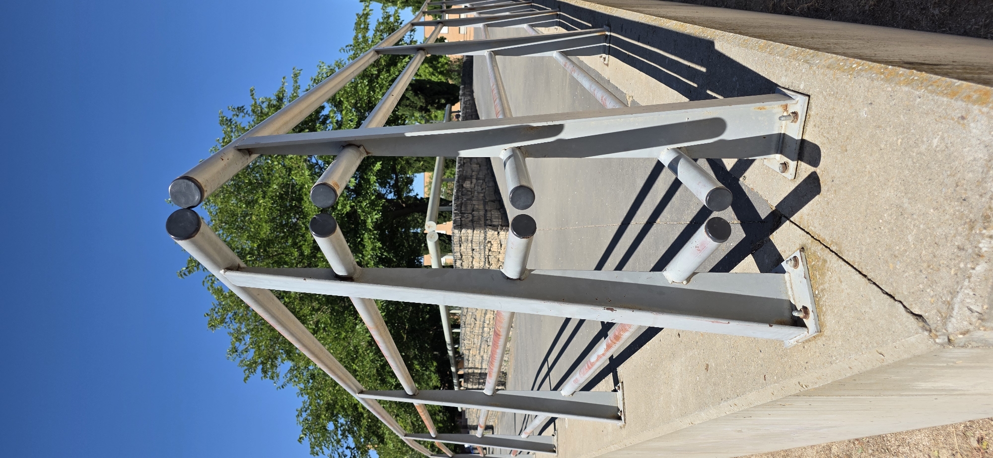 The image shows a metal railing with a grey finish and some rust spots, attached to a concrete base. The railing is positioned on a sidewalk or pathway, with trees and a blue sky in the background. The sun is casting shadows from the railing onto the pavement. The railing appears to be part of a park or public space. Based on the architectural style of the stone wall in the background and the trees, it is likely a public area in the city of Huesca, Spain. No people are visible in the frame.
