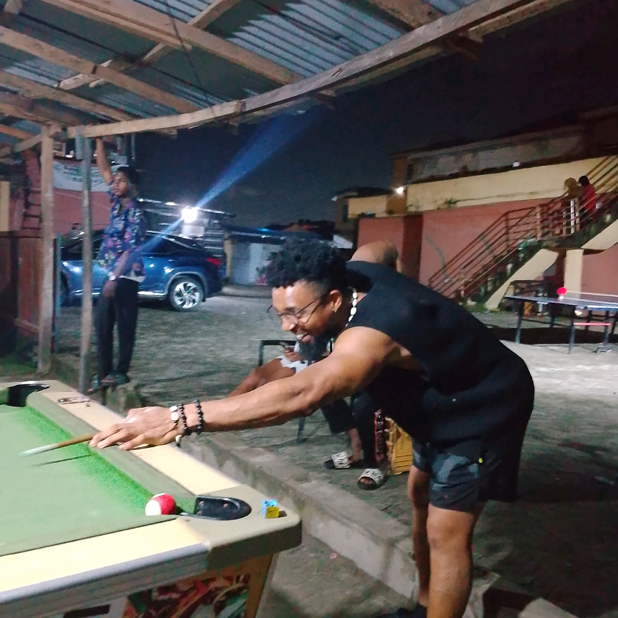 In Ado, Nigeria, a man is playing pool at night. He is wearing a black t-shirt and shorts and is in the process of striking the cue ball. A red and white billiard ball is near the pocket. A blue car is parked behind him. In the background, a person is visible on a staircase. The scene is outdoors, under a wooden structure with a metal roof.