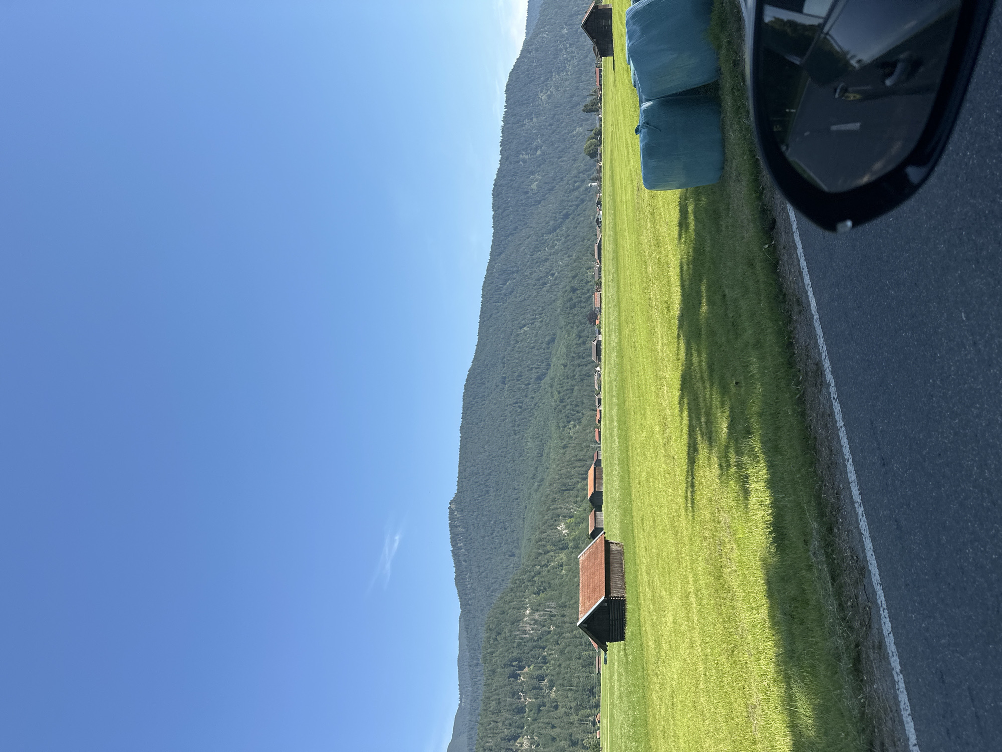 The image depicts a scenic view of a green field alongside a road, with a densely forested mountain range in the background. Several small wooden structures with red roofs are scattered across the field, and large, blue-wrapped hay bales are visible on the right side of the frame. A car's side mirror, reflecting the sky and surrounding landscape, is prominent in the foreground on the right. The scene is illuminated by bright sunlight, suggesting a clear and sunny day. The clear blue sky indicates good weather. The presence of the hay bales and the rural setting suggests an agricultural area. The image appears to be taken from a moving vehicle, given the perspective and the car mirror. The landscape, with its mountains and traditional buildings, points to a European Alpine region. The location context provided is Garmisch-Partenkirchen, Germany, which aligns with the visual cues. No people are visible in the image. The overall mood is serene and picturesque, highlighting the natural beauty of the countryside. There is no visible text in the image.