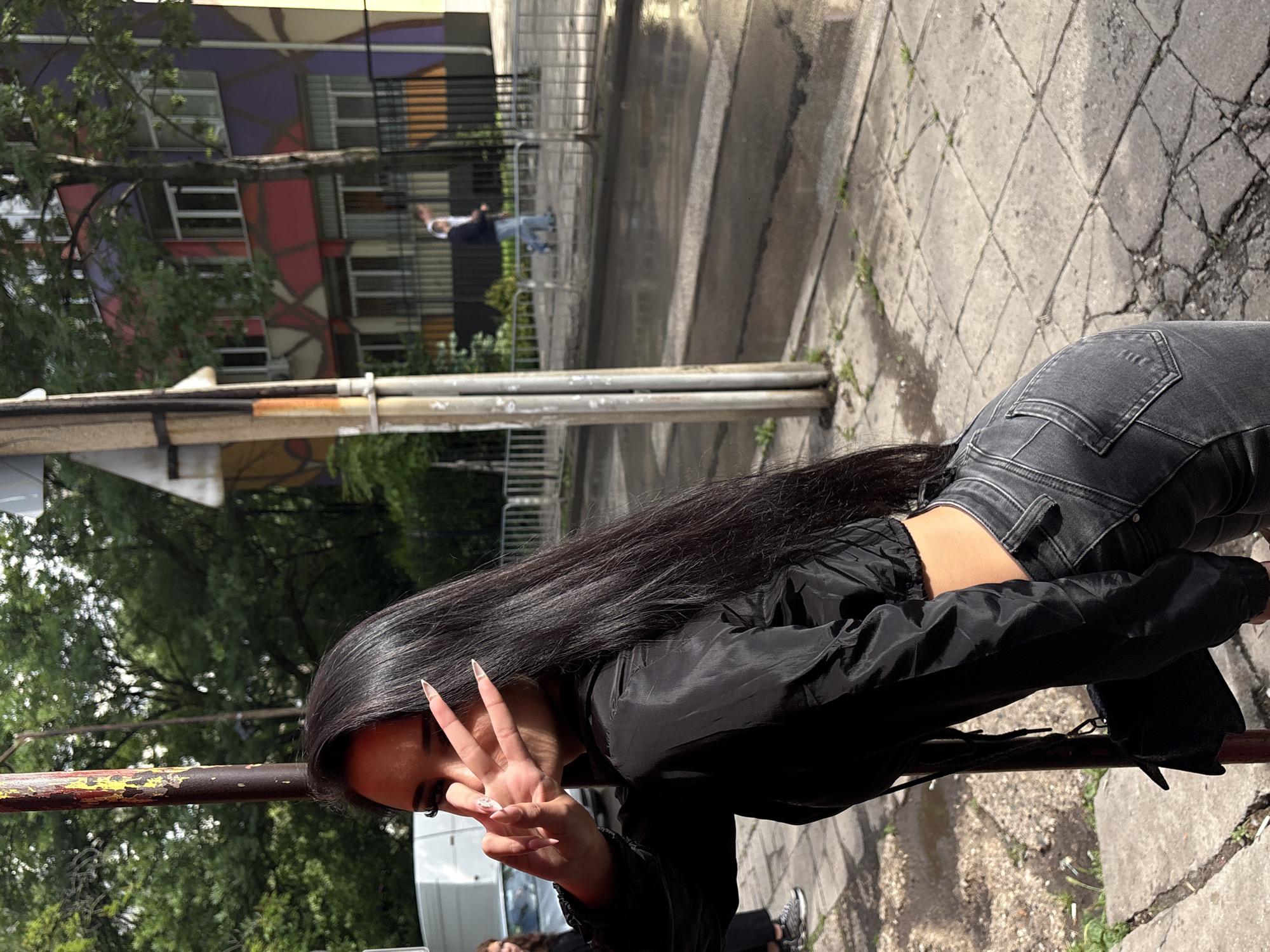 The image displays an outdoor scene in what appears to be an urban environment in Sofia, Bulgaria, under bright daylight. The primary subject is a person viewed from behind and slightly to the side, with long, straight dark hair. They are dressed in a black long-sleeved top or jacket and dark grey jeans, with a small portion of their lower back visible between the garments. The person is holding up their left hand, making a peace sign with noticeably long, pointed, light-colored nails.
A rusty metal railing with hints of yellow and red paint runs diagonally across the mid-ground, positioned in front of the person. The ground consists of concrete paving stones, showing cracks and some small patches of green vegetation.
In the background, lush green trees and foliage are visible. Beyond them, a multi-story building with a striking, abstract, geometric facade in shades of purple, red, and yellow/gold stands out. A paved street with crosswalk markings is also discernible, and faint outlines of distant people, including one near a white van, can be seen further back. Shadows suggest a sunny day. No visible text is present in the image.
