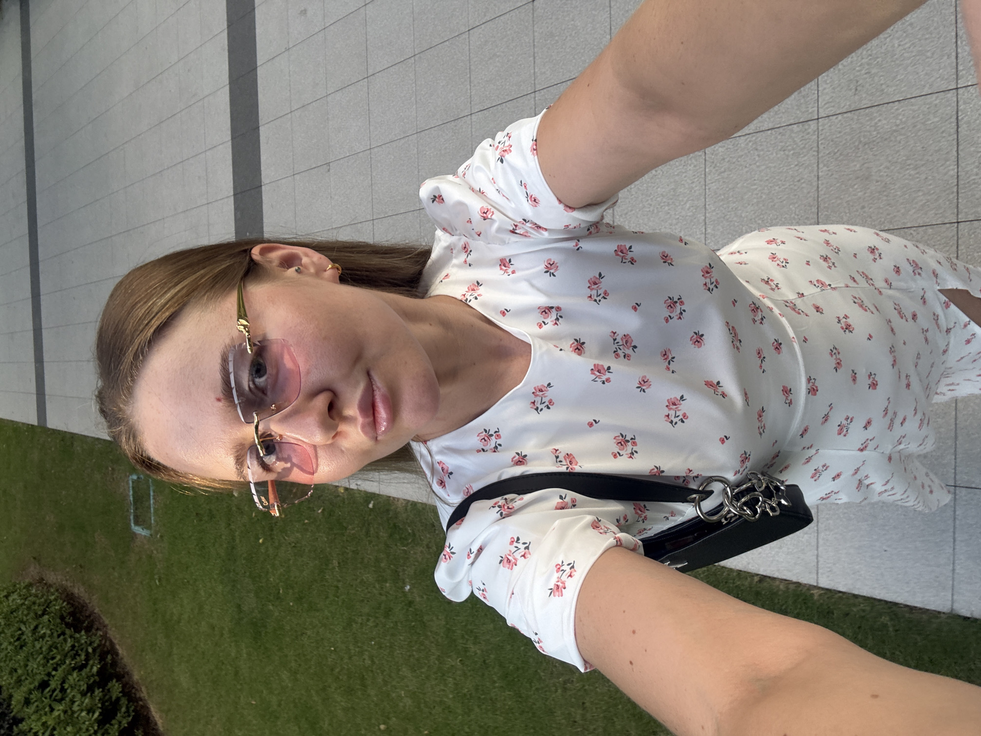 A medium shot captures a young woman from the chest up, taking a selfie outdoors. She has blonde hair and is wearing pink-tinted, gold-rimmed glasses and a white dress adorned with a floral pattern of pink roses. A black handbag hangs from her left shoulder. The background is a mix of green grass and a paved walkway composed of rectangular grey tiles. The woman is positioned slightly off-center to the right.