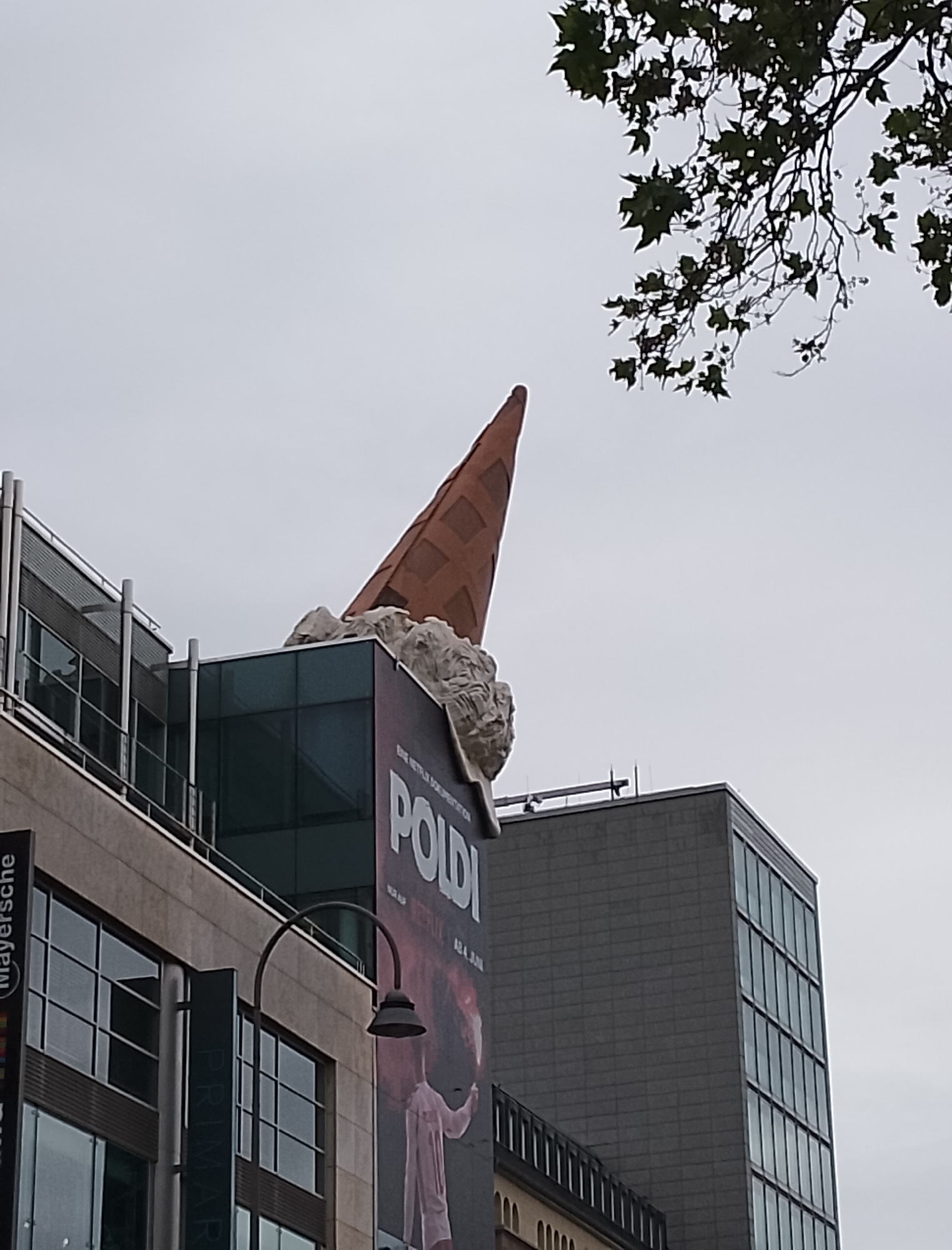 The image shows a large, orange ice cream cone sculpture atop a building. The cone appears to be made of a waffle material and is pointing upwards at an angle. Below the cone, there is a white, textured object that resembles melting ice cream. The sculpture is positioned on a building with modern architecture, featuring glass and metal elements. A large poster is visible on the side of the building, featuring the word "POLDI" in bold white letters and a depiction of a person in a light-colored suit. The poster also mentions "BNE NETFLIX DOCUMENTATION". To the left of the sculpture, another building with large windows and balconies is visible. In the upper right corner, a tree with green leaves is partially obscuring the cloudy sky. The overall scene suggests an urban setting with an artistic installation. The lighting indicates it is daytime, and the overcast sky suggests cloudy weather. No specific emotions are conveyed, but the art piece adds an element of whimsy to the urban landscape. Visible text includes "Meyersche", "PRIMARK", and "POLDI".