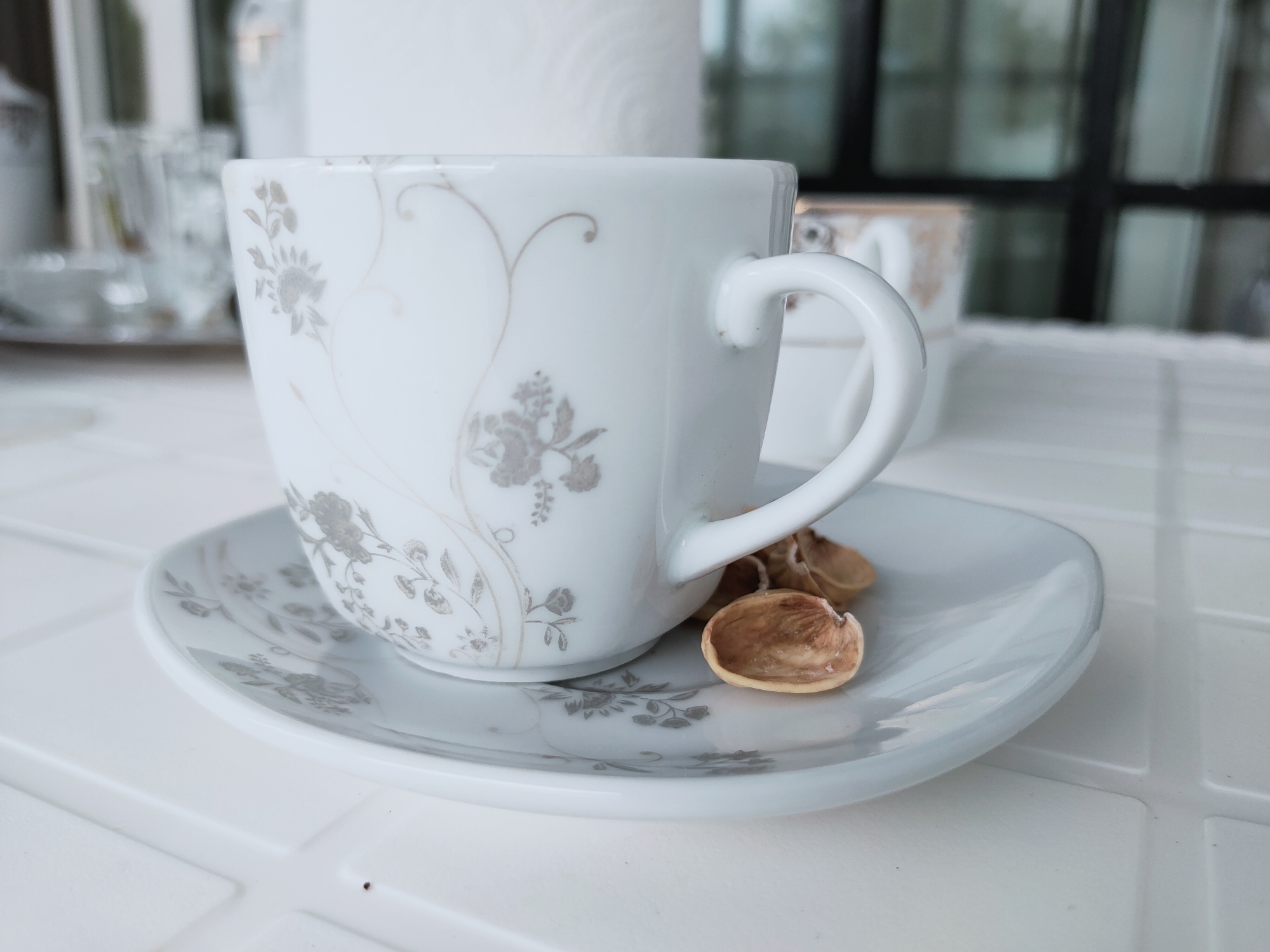 A white teacup with a floral pattern sits on a matching saucer. Next to the teacup on the saucer are two dry, light brown almond shells. The teacup and saucer are on a white surface with a grid pattern. In the background, another teacup is partially visible. The overall scene appears to be set indoors, potentially on a table in a home or cafe in Mrezga, Tunisia, given the lighting and style of the tableware.