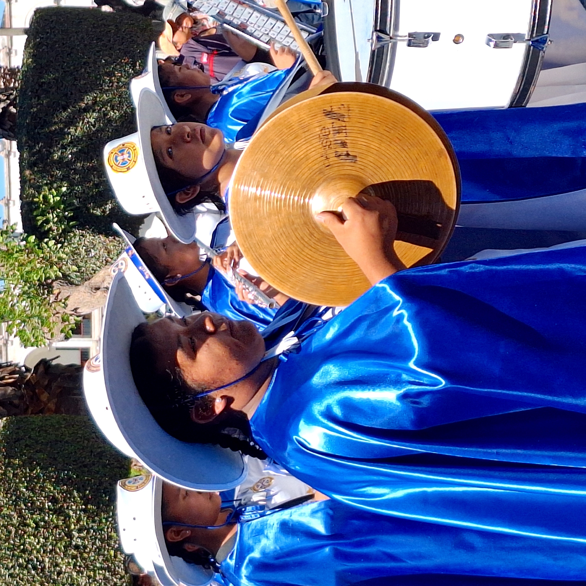 In the city of Unknown, Unknown, a group of young people participate in a parade or performance. They are dressed in matching blue gowns and white hats with a symbol on the front, suggesting they are part of a marching band or a similar organization.
One individual is holding and playing a large, golden cymbal. Another appears to be playing a xylophone, while others may be playing flutes or other instruments. There is a large white drum visible as well.
The environment includes trees and possibly buildings in the background, indicating the event is taking place outdoors in a public space. The overall scene suggests a community event or celebration.