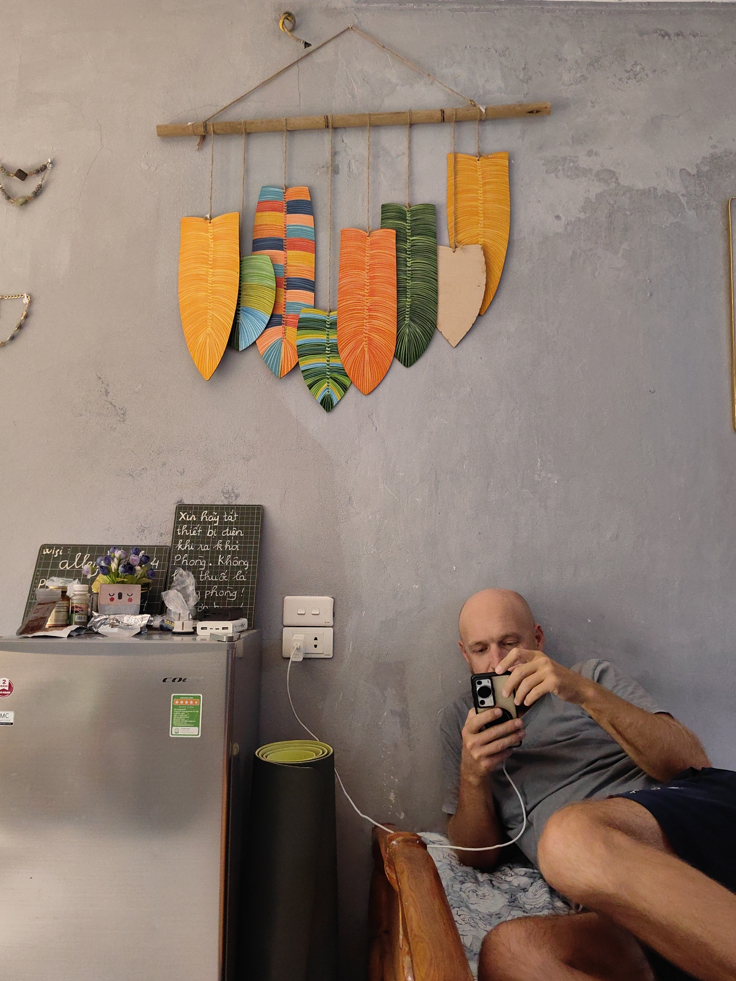The image depicts a bald man lying on a bed, intently focused on his smartphone. He wears a gray t-shirt and dark blue shorts. Behind him is a grey concrete wall adorned with a decorative bamboo and fabric art piece with a leaf motif. To the left of the man is a stainless steel refrigerator with a green energy efficiency sticker and various items piled on top, including bottles, chargers, and a small chalkboard with writing in Vietnamese. A rolled black yoga mat stands beside the refrigerator. The scene appears to be indoors in a small living space and is consistent with a residence or a hotel room in Hà Nội, Vietnam, based on the visible text and general appearance.