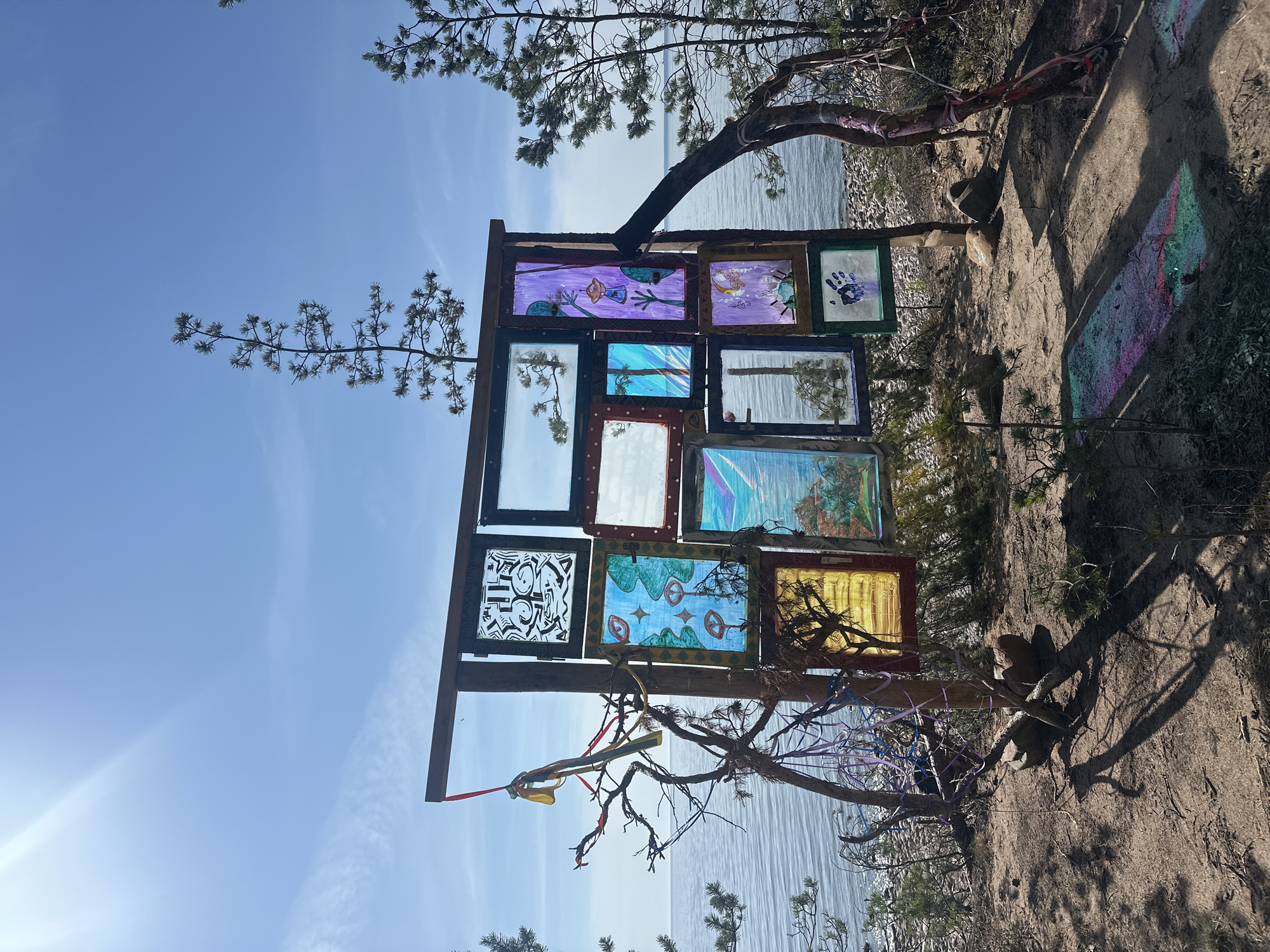 This image depicts an outdoor art installation located on a sandy shore in Luzhki, Russia, with a body of water in the background and trees on either side of the frame. The installation is a wooden structure with multiple framed panels, some of which appear to be made of glass or transparent material with artwork or drawings on them. The artwork includes various abstract and representational images, such as faces, trees, and handprints, rendered in different colors and styles. Some panels are clear, reflecting the sky and surrounding trees. The structure is supported by wooden posts and is adorned with colorful ribbons and string, adding to its whimsical appearance. In the foreground, the sandy ground is scattered with small stones and sparse vegetation. The sky is bright blue with a few wispy clouds, and sunlight casts shadows across the scene. The overall atmosphere is serene and artistic, suggesting a creative expression within a natural landscape.