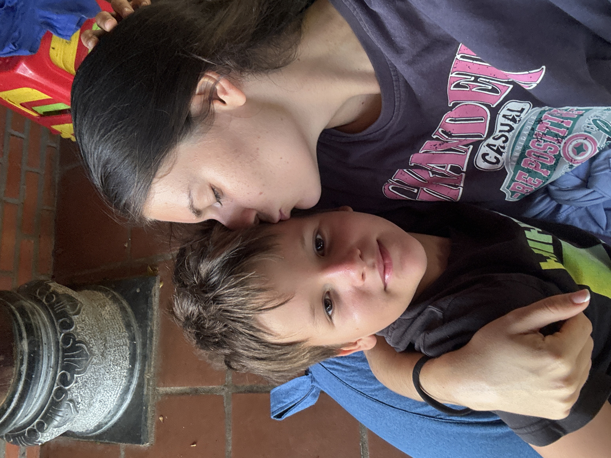In Mũi Né, Vietnam, a young woman with dark hair is kissing a young boy on the forehead. She is wearing a dark t-shirt with a graphic design. The boy is wearing a dark t-shirt with a lime green graphic. The boy is sitting on a blue cushion. The woman is wearing a black hair tie on her wrist. In the background, there is a stone pillar with decorative carvings and reddish-brown brickwork. A red and yellow plastic toy is also visible in the background. The scene appears to be outdoors.