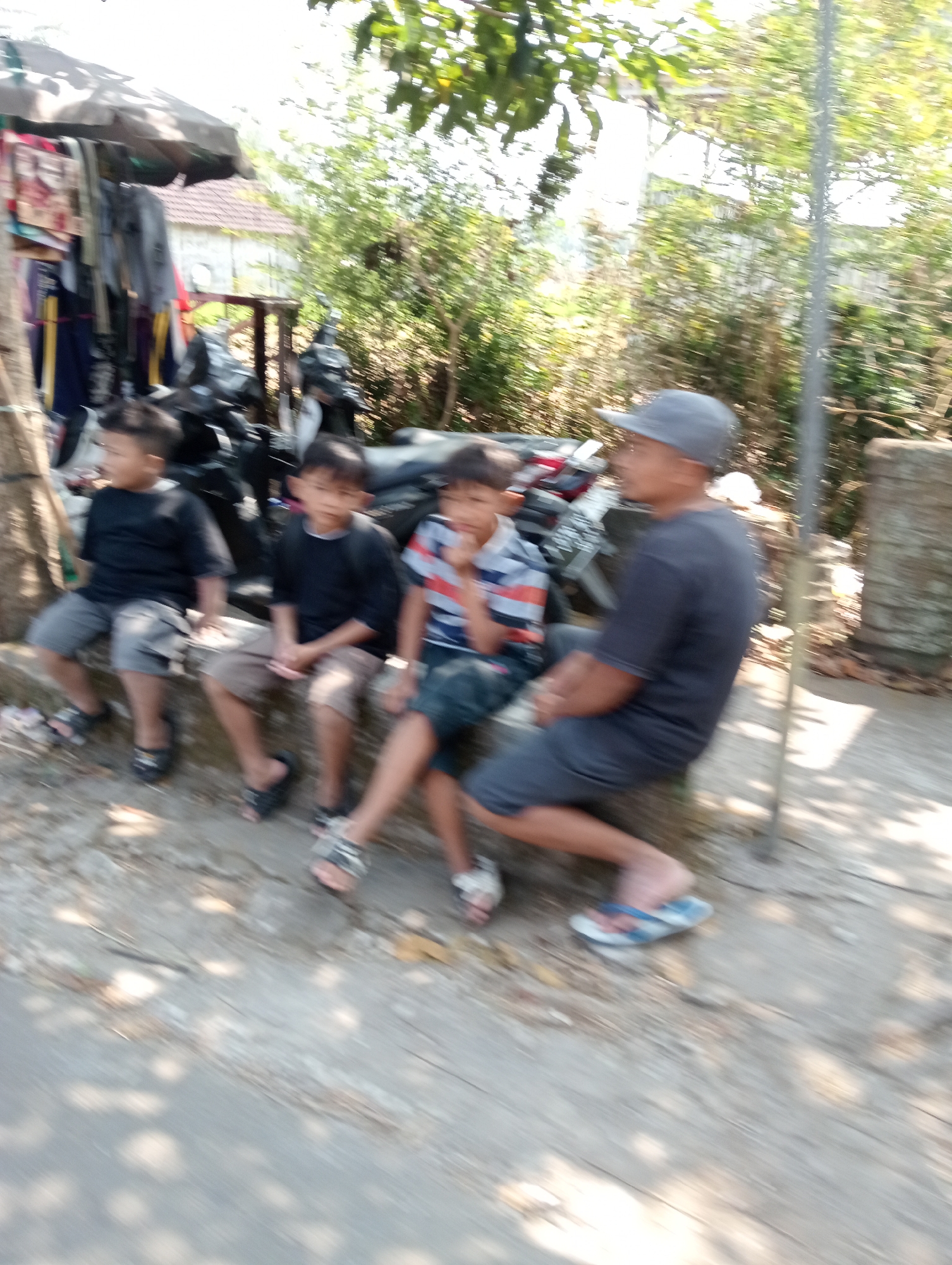 In the city of Krajan Kidul, Indonesia, on a sunny day, four individuals are seated on a concrete ledge. On the far left, a young boy, wearing a dark t-shirt and grey shorts, is seen from the side, looking to his left. Next to him, another young boy, also in a dark t-shirt and grey shorts, faces forward. The third person, a young boy in a striped t-shirt and dark shorts, sits with his legs crossed and is looking downwards. To the right, an adult male wearing a grey cap and a dark t-shirt is seated, facing to his left. Behind the individuals, several motorcycles are parked. The background consists of lush green trees and foliage, indicating an outdoor setting, possibly near a street or path.