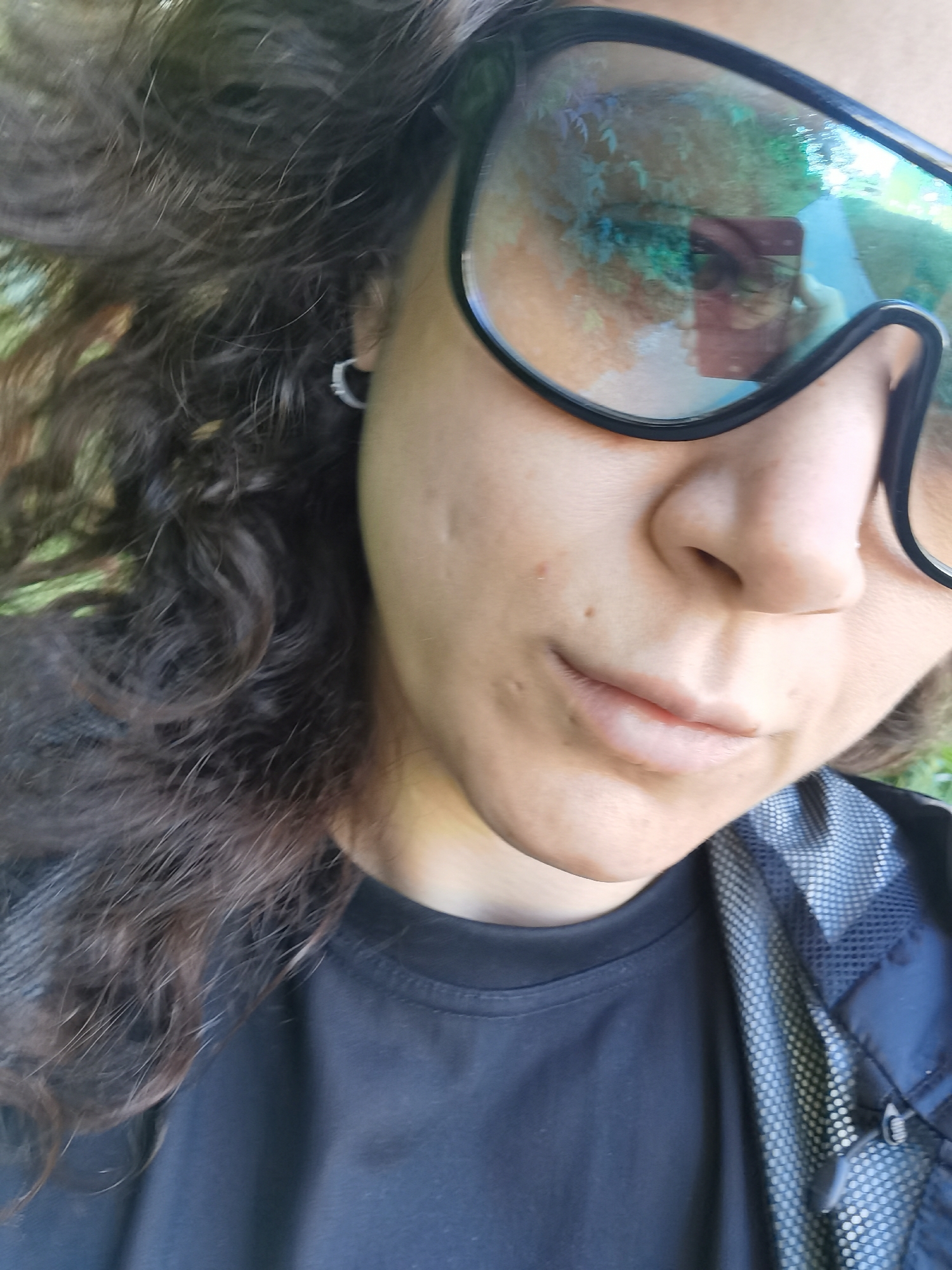 The image is a close-up, eye-level, over-the-shoulder selfie of a person wearing large, black-framed goggles with reflective lenses. The person has dark, curly hair that is blowing in the wind. They are wearing a black t-shirt, and the collar of a dark jacket with a light-colored mesh lining is visible. The background is out of focus and appears to be an outdoor environment with green foliage, suggesting a park or natural setting. The reflection in the goggles shows a mobile phone being held by the person, indicating they are taking the photo. There are no discernible landmarks or features that would place this image specifically in the city of Chaikovsky, Russia.