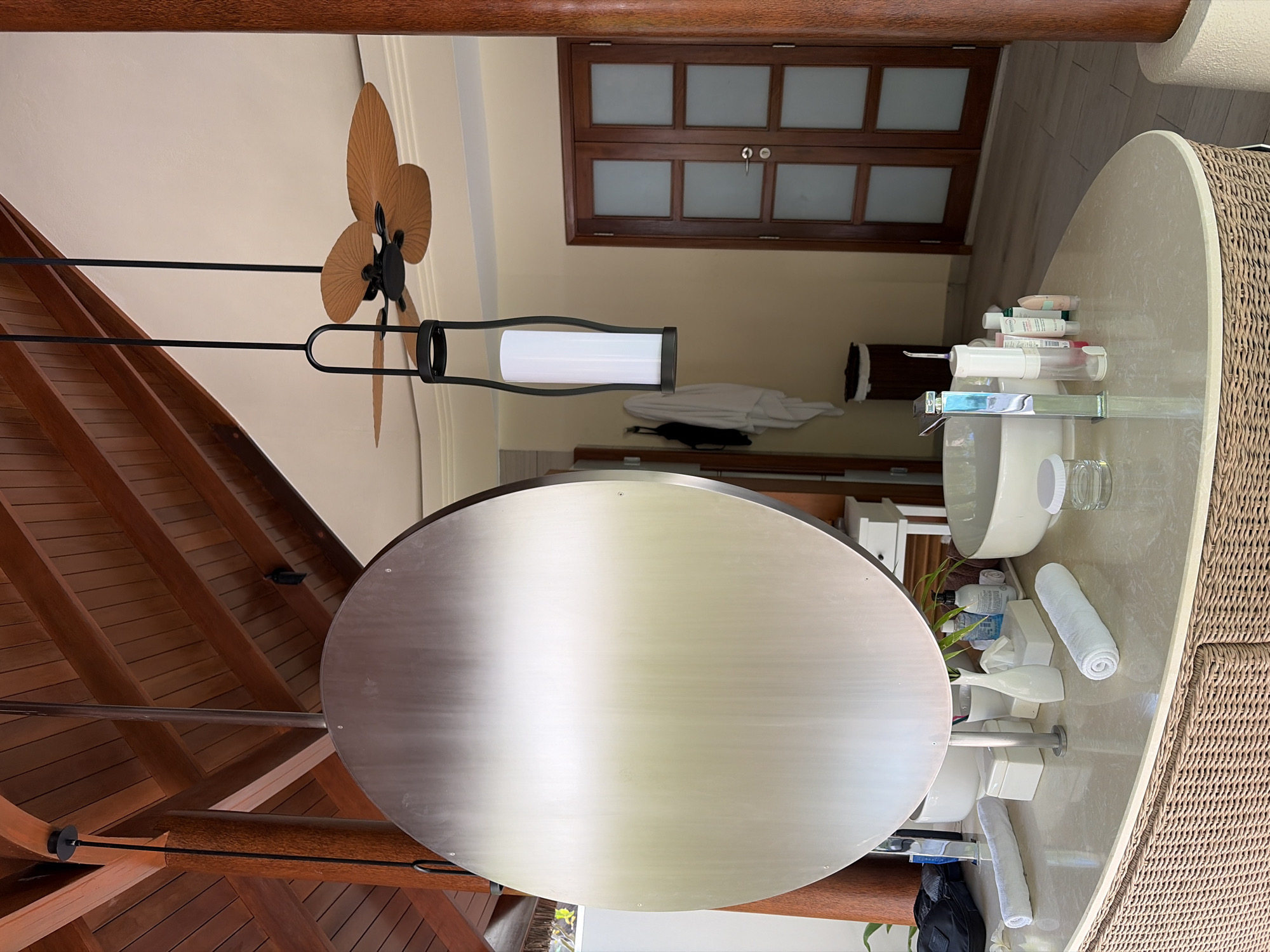 The image shows a bathroom area in Halaveli, Maldives. A large, round, brushed metal mirror hangs prominently in the foreground. To the right, a ceiling fan with wooden blades is visible, along with a modern hanging lamp. A vanity countertop, made of light-colored stone and supported by a wicker base, holds various toiletries, including bottles of lotion, tissue boxes, rolled towels, and a sink. In the background, wooden double doors with frosted glass panels are visible, suggesting an adjoining room or exit. The architecture features exposed wooden beams on the ceiling, contributing to a tropical, resort-like aesthetic. There are no people present in the scene.