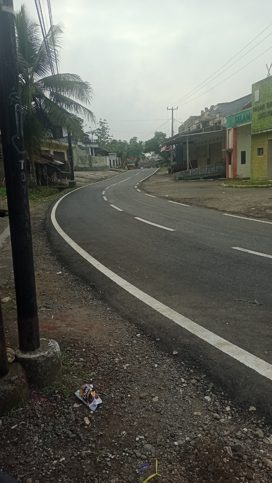 The image shows a curved asphalt road in Cogreg, Indonesia, with white dashed lines marking the lanes and a solid white line along the edge. The road passes by several buildings with corrugated metal roofs on the right side, one of which has a green facade with red and white trim and a sign that appears to read "SELAMAT DATANG" (Welcome). Further down the road, a red and yellow truck is visible. On the left side of the road, there is a dark pole with some graffiti, a palm tree, and some small structures with weathered roofs. The ground is covered with gravel and dirt. There are some discarded wrappers and debris on the side of the road. The sky is overcast and the lighting suggests it is daytime. No people are clearly visible in the image.