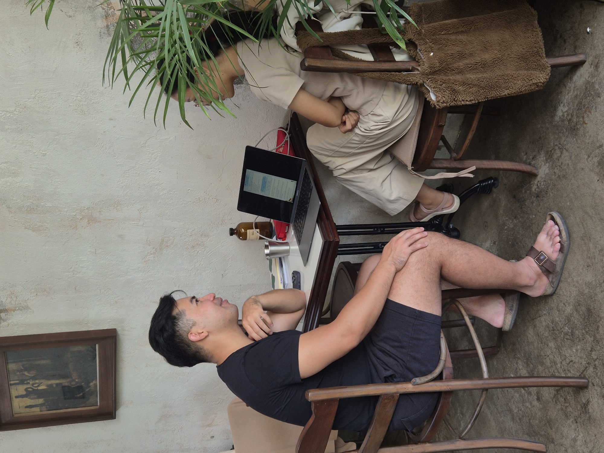 A young man with dark hair, wearing a black t-shirt and black shorts, sits on a wooden chair to the left of a small table. He is wearing brown sandals. He is looking towards a young woman sitting on a wooden chair to his right. The woman has dark hair and is wearing a light-colored t-shirt and light-colored pants. She is wearing pink sandals. She is looking at a laptop placed on the table. On the table are a bottle, a metal cup, a phone, and some papers. A framed painting hangs on the textured wall behind the man. A large brown furry bag hangs on the back of the woman's chair. Plants are visible in the foreground, obscuring part of the woman. The scene appears to be an indoor cafe, possibly in Đà Nẵng, Vietnam.
