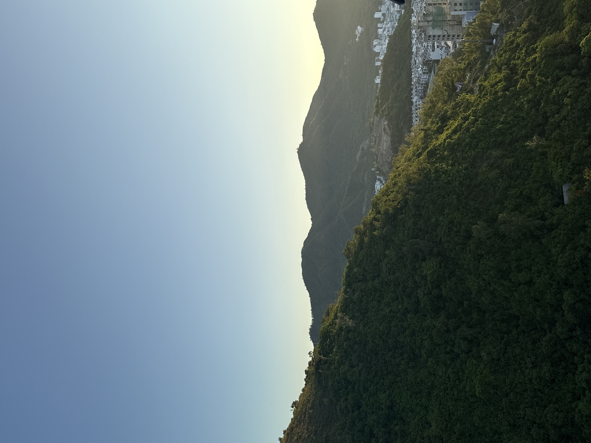 This image captures a sweeping view of the landscape surrounding Nha Trang, Vietnam, likely taken from an elevated vantage point overlooking the city. In the foreground, a densely forested, steep mountainside dominates the left half of the frame, its lush green foliage bathed in the warm glow of what appears to be late afternoon or early morning sunlight.
In the mid-ground, the forest gives way to a sprawling urban development. Rows of white and light-colored buildings, characteristic of residential areas, are visible nestled among the hills. Some buildings appear to be under construction, indicated by exposed concrete structures and scaffolding, suggesting ongoing development in the city.
Further in the background, a series of undulating, dark green mountains rise, their silhouettes softened by distance and the atmospheric haze. The sky above is a clear, gradient of pale blue, transitioning to a lighter, almost yellowish hue near the horizon where the sun is presumably located. The overall impression is one of a coastal city coexisting with its natural, hilly surroundings. No people or specific activities are discernible in this particular shot.